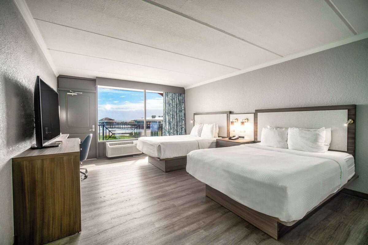 A hotel room with two beds, a desk with a TV, a large window with a city view, and a balcony.