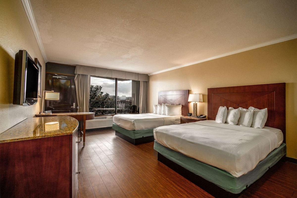 A hotel room with two beds, a desk, a TV, a balcony with a view, and a large window.