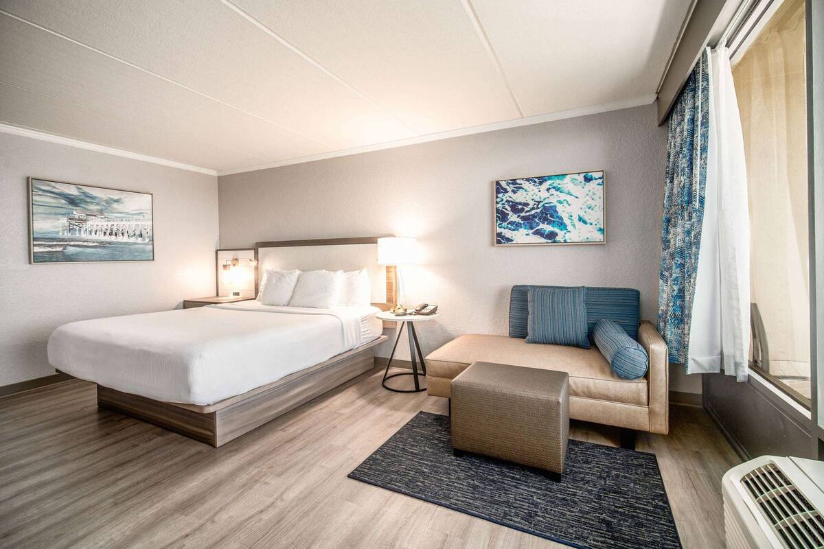 A hotel room with a bed, a sofa, a small table with a lamp, and artwork on the walls.