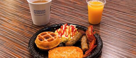 Plato de desayuno con waffles, huevos revueltos, salchichas y una guarnición de patatas ralladas sobre un plato negro, acompañado de una taza de café y un vaso de zumo de naranja sobre una mesa a rayas.