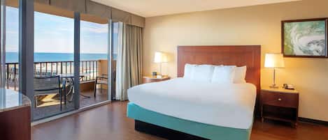 A hotel room with a large bed, two bedside tables with lamps, a view of the sea through a sliding glass door, and a balcony with outdoor seating.