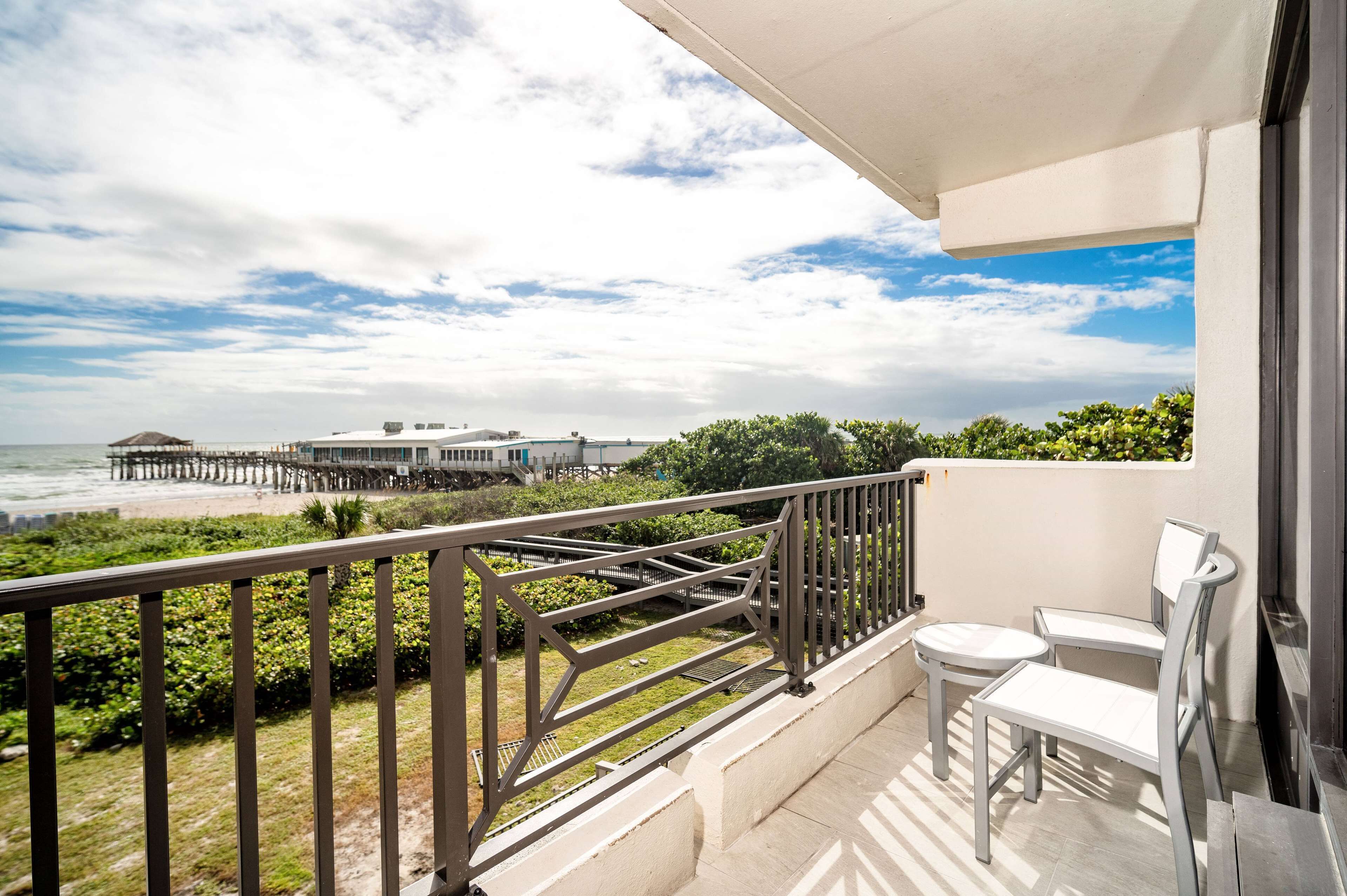 A balcony with a view of the beach and pier, two white chairs, and a railing.