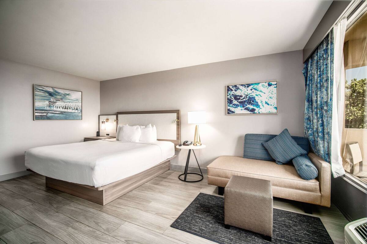 A modern hotel room with a large bed, a sofa, a small table with a lamp, and artwork on the walls.