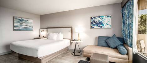 A modern hotel room with a large bed, a sofa, a small table with a lamp, and artwork on the walls.