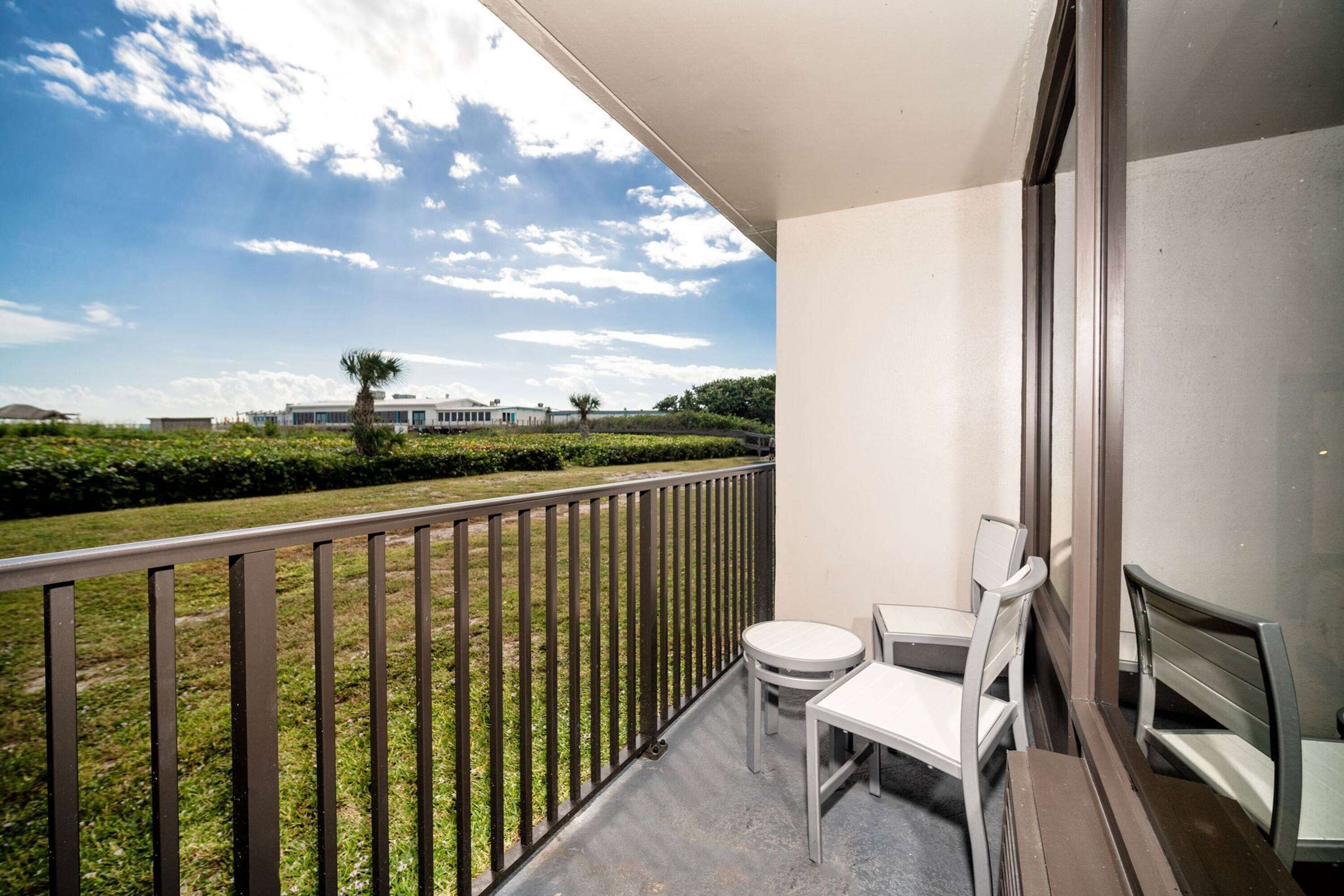 A balcony with a view of a landscaped garden and a building in the distance. There are two white chairs and a small table on the balcony.