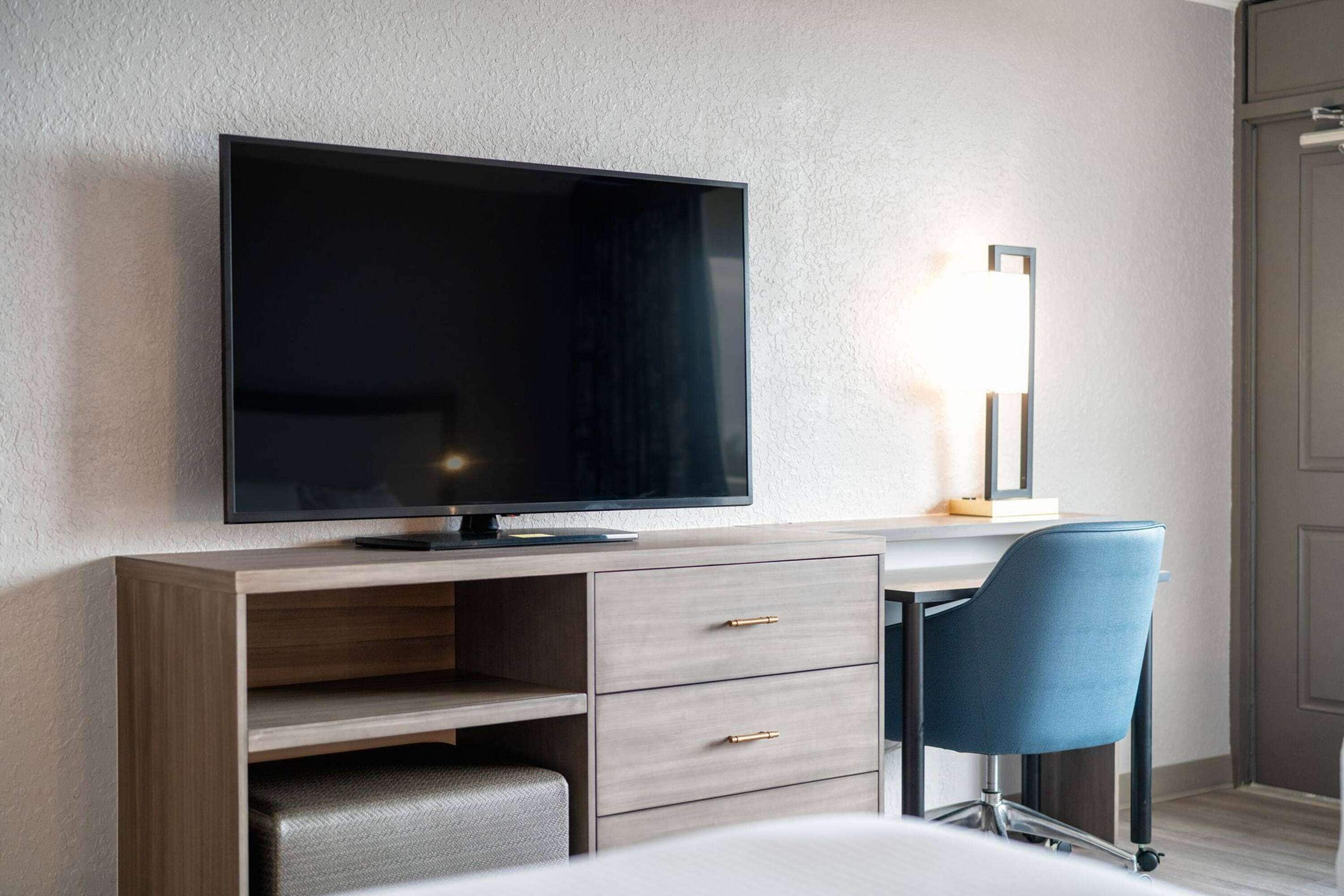 Habitació d'hotel moderna amb televisió de pantalla plana penjada sobre un escriptori de fusta, una cadira blava i una taula de nit.