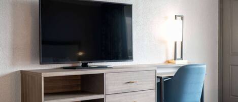 A modern hotel room with a flat-screen TV mounted on a wooden desk, a blue chair, and a bedside table.