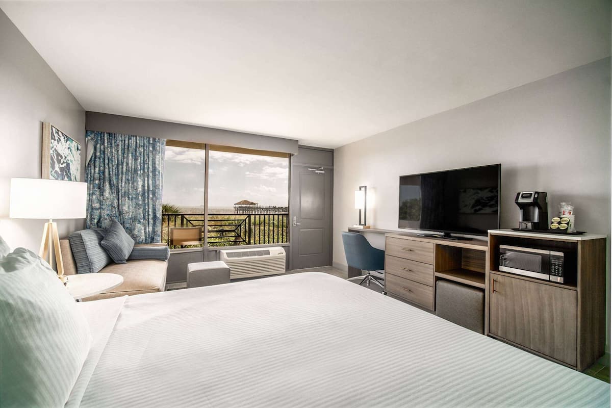A modern hotel room with a large bed, a desk with a flat-screen TV, a microwave, and a coffee maker. There is a balcony with a view of greenery.