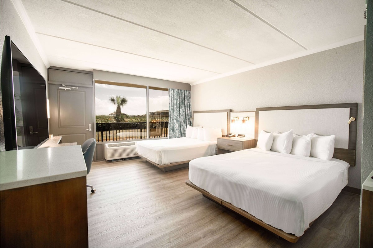 A hotel room with a large bed, a desk, a TV, and a balcony with a view of palm trees.