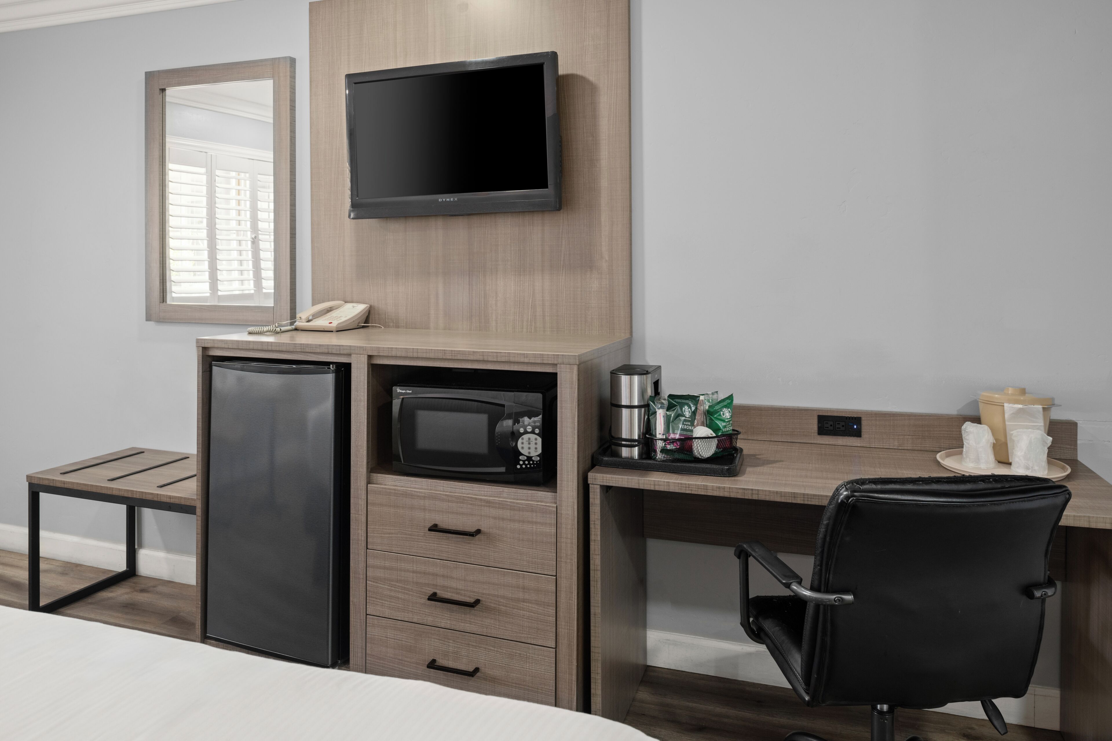A hotel room with a bed, a desk with a chair, a television, a microwave, a refrigerator, a coffee maker, and a telephone.