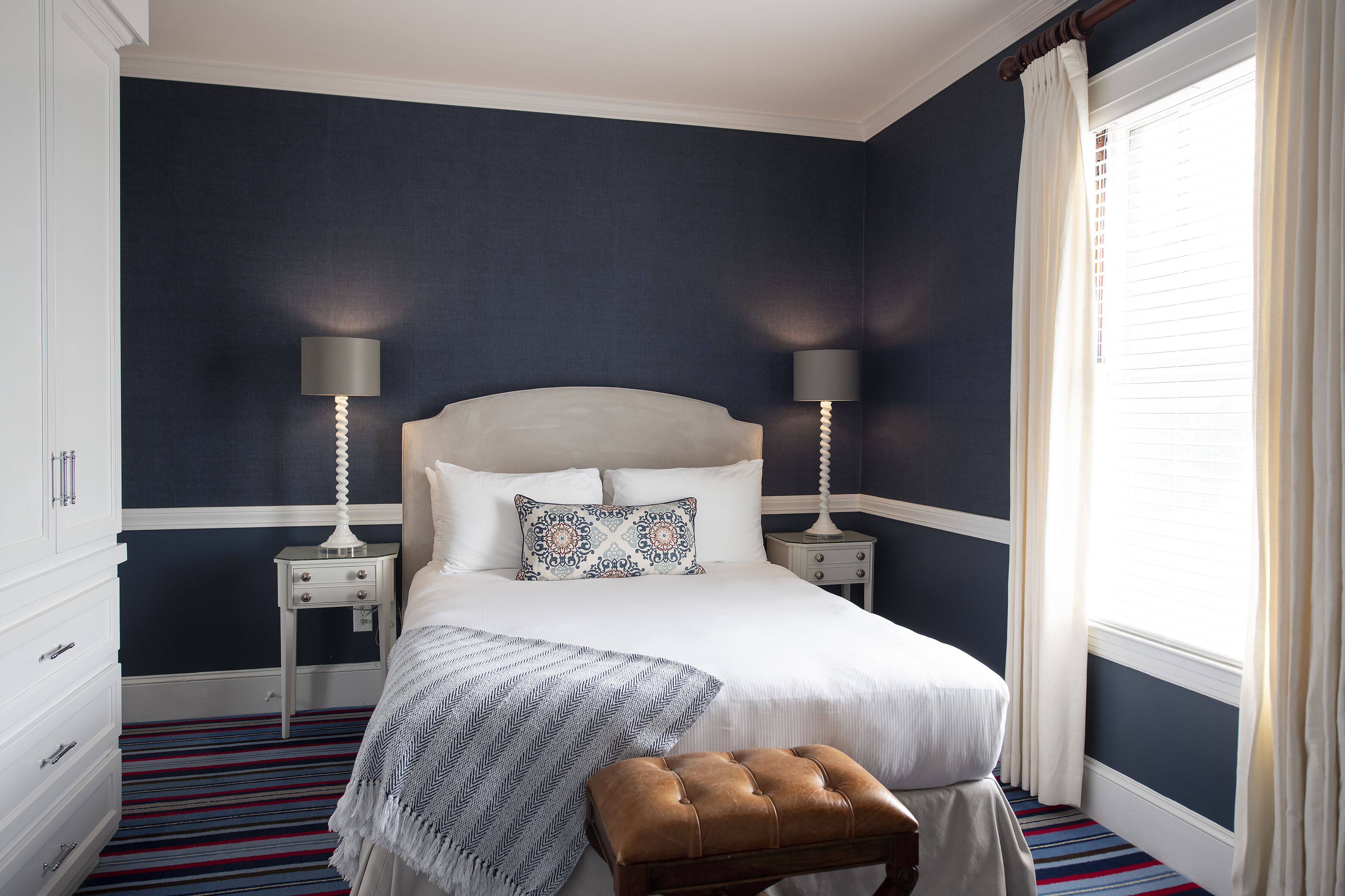 A bedroom with a large bed, two bedside tables, a nightstand, a bench, and a window with curtains.