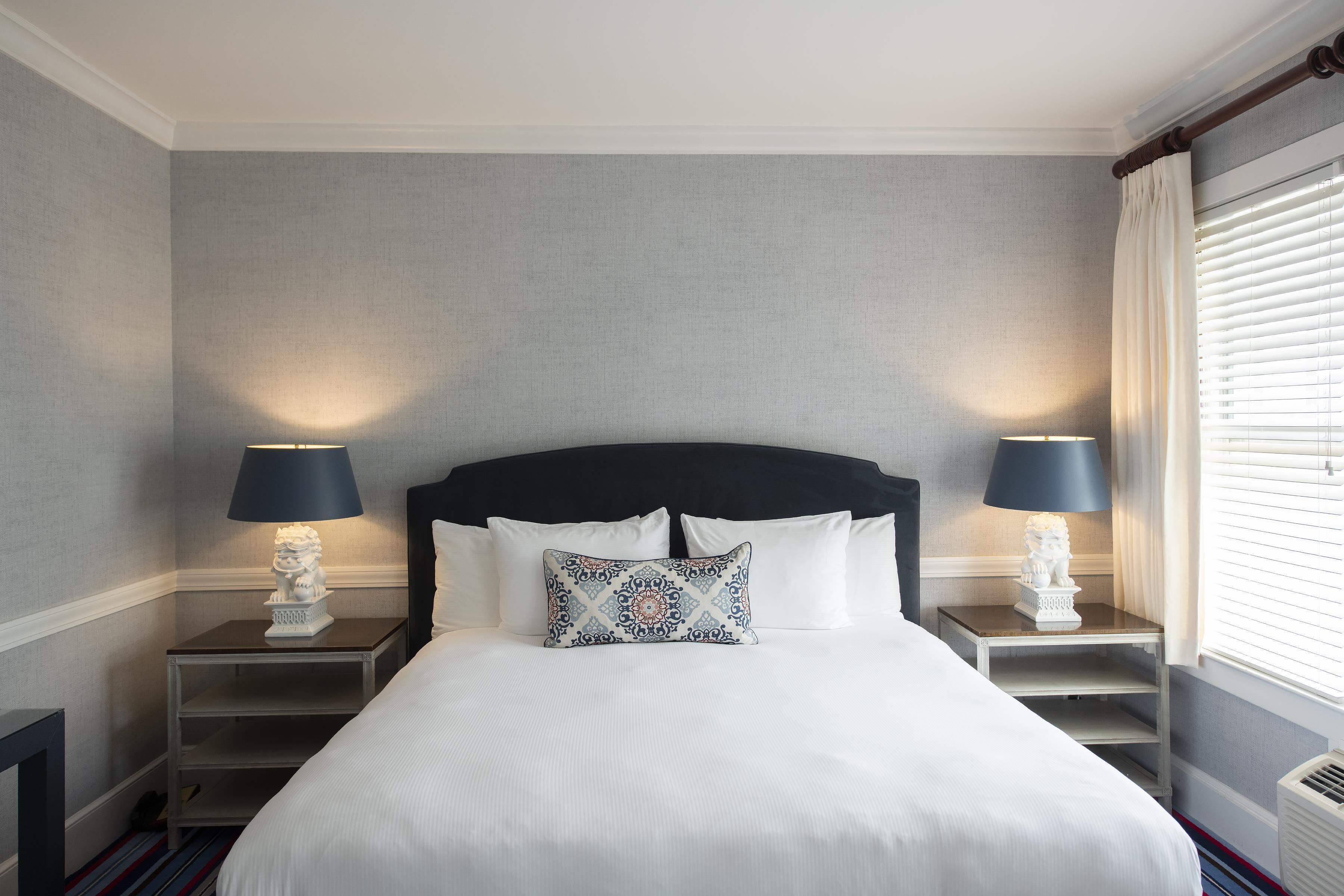 A neatly made bed with white bedding, two bedside tables with lamps, and a window with blinds.