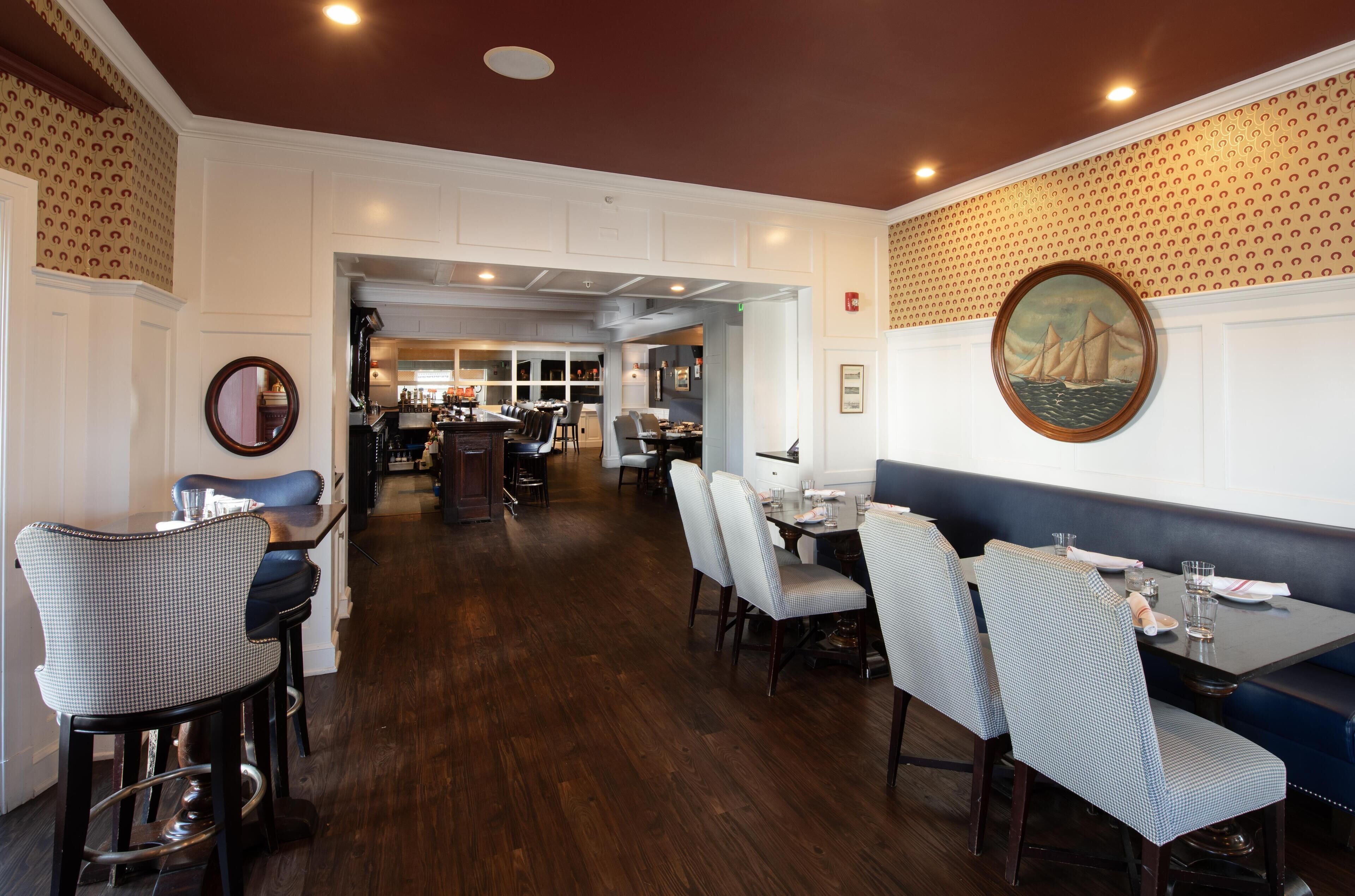 Un restaurante con suelo de madera, paredes blancas y papel tapiz estampado. Se observan mesas y sillas de comedor, y una zona de cocina al fondo.