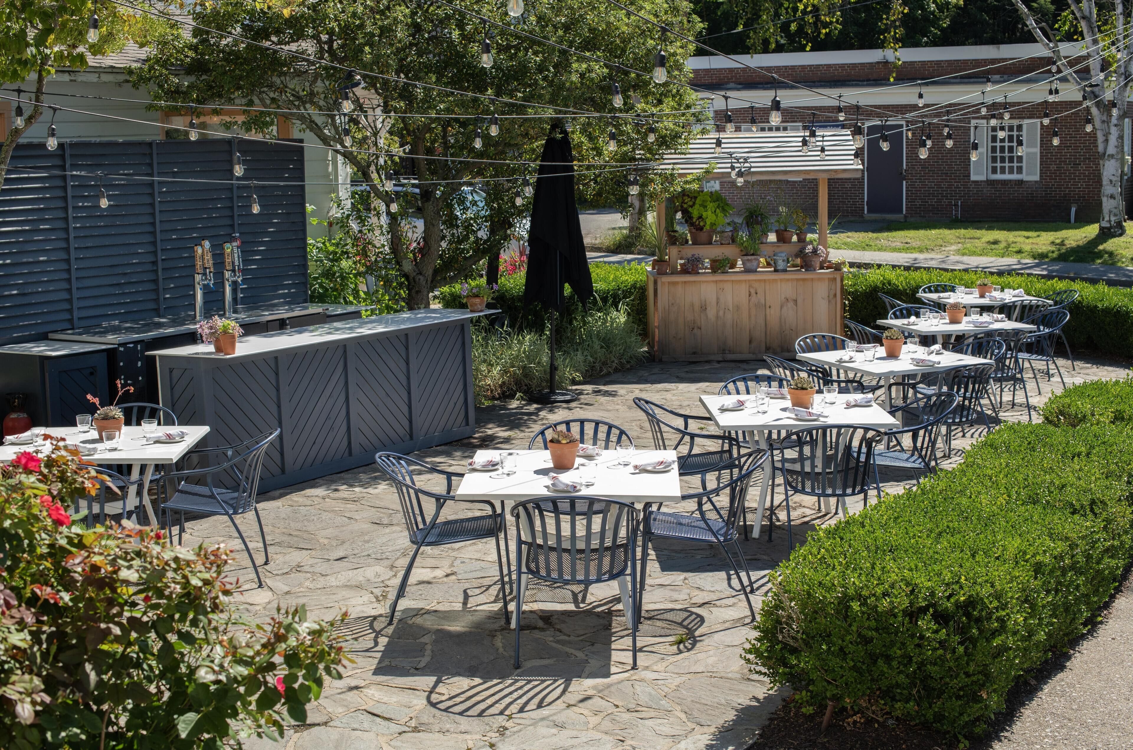 Een terras buiten met tafels en stoelen, een bar en potplanten.