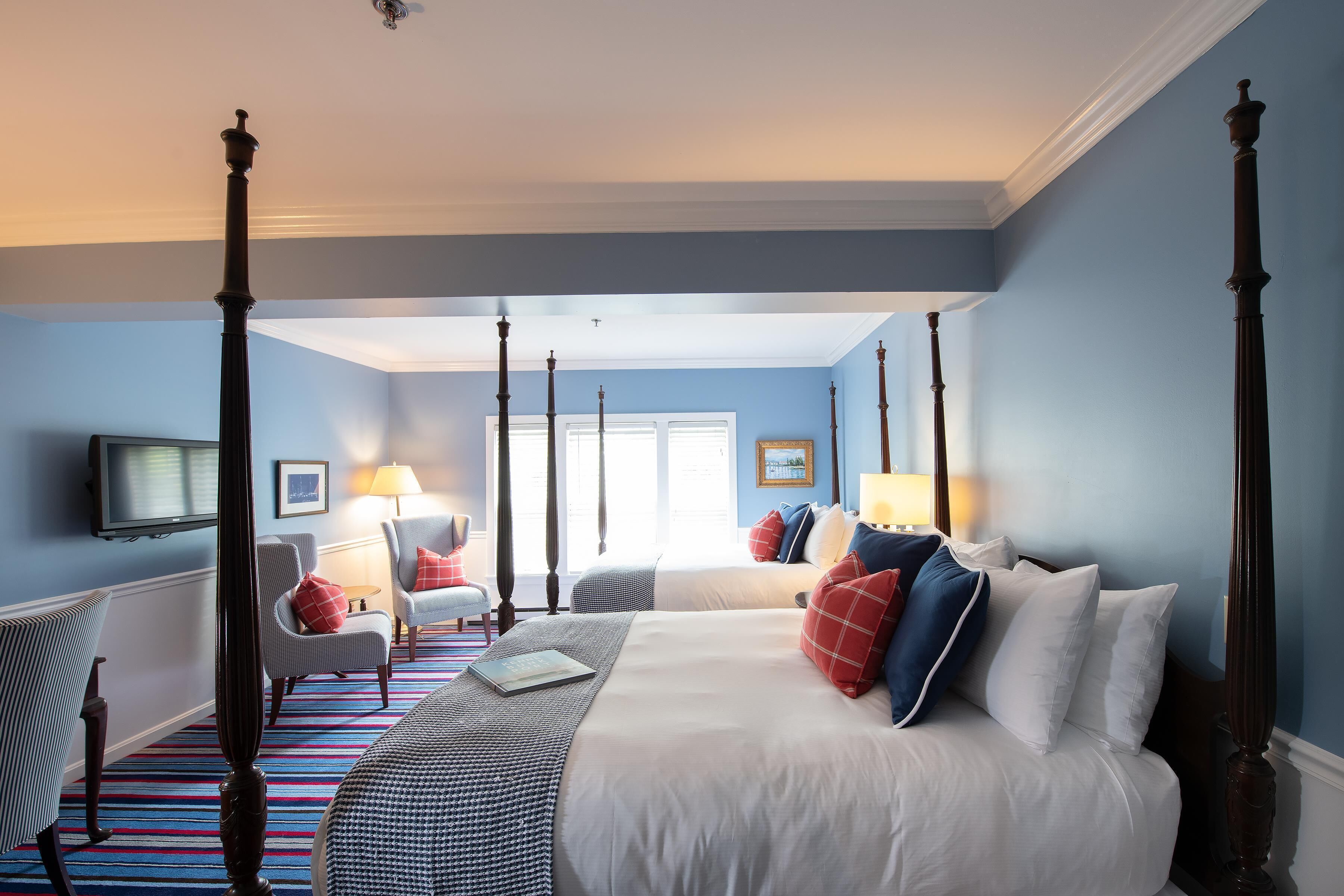 A four-poster bed with white bedding, a striped carpet, and a view of a window with a lamp and chair in the background.