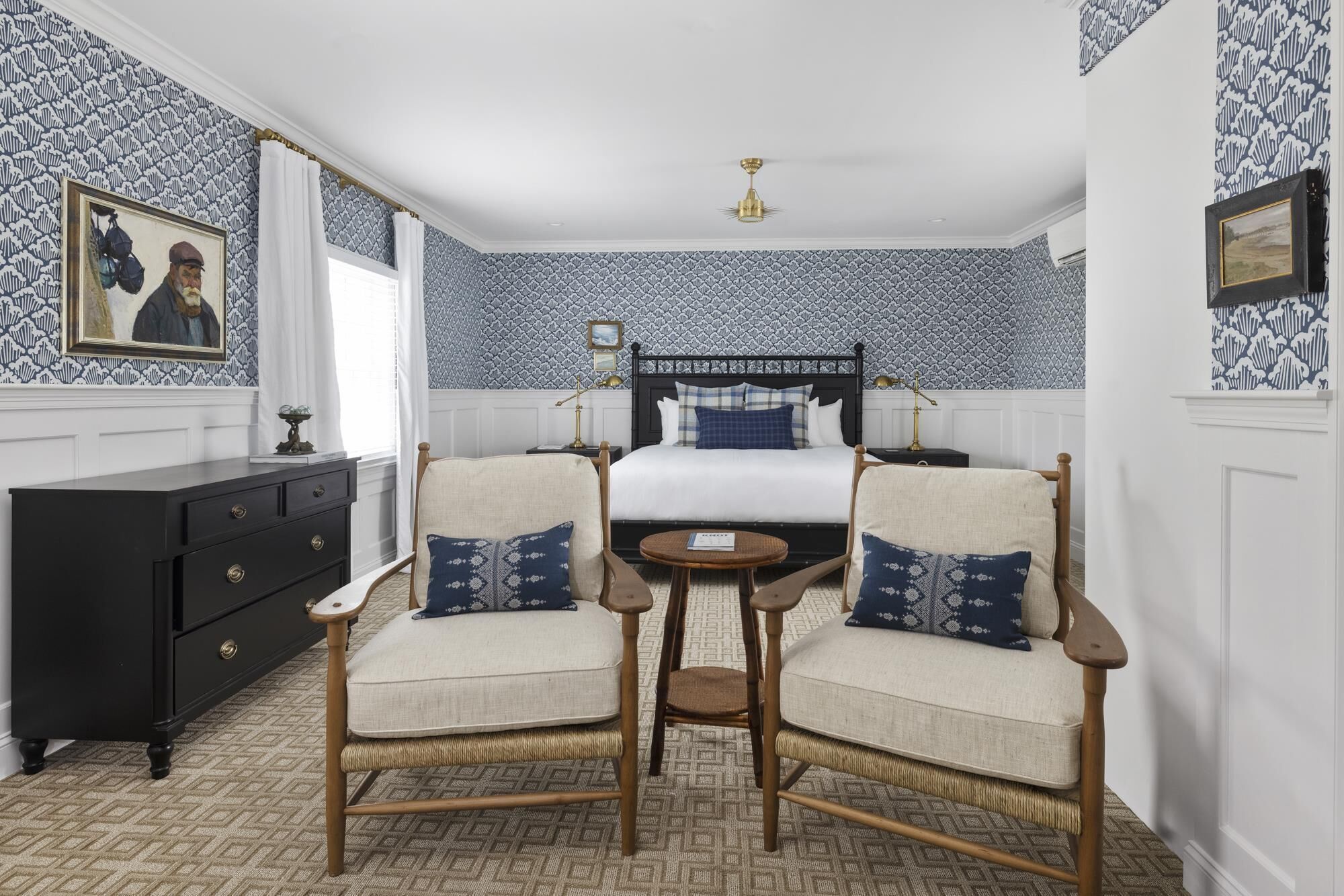 A hotel room with a bed, two armchairs, a small table, a dresser, and patterned wallpaper.