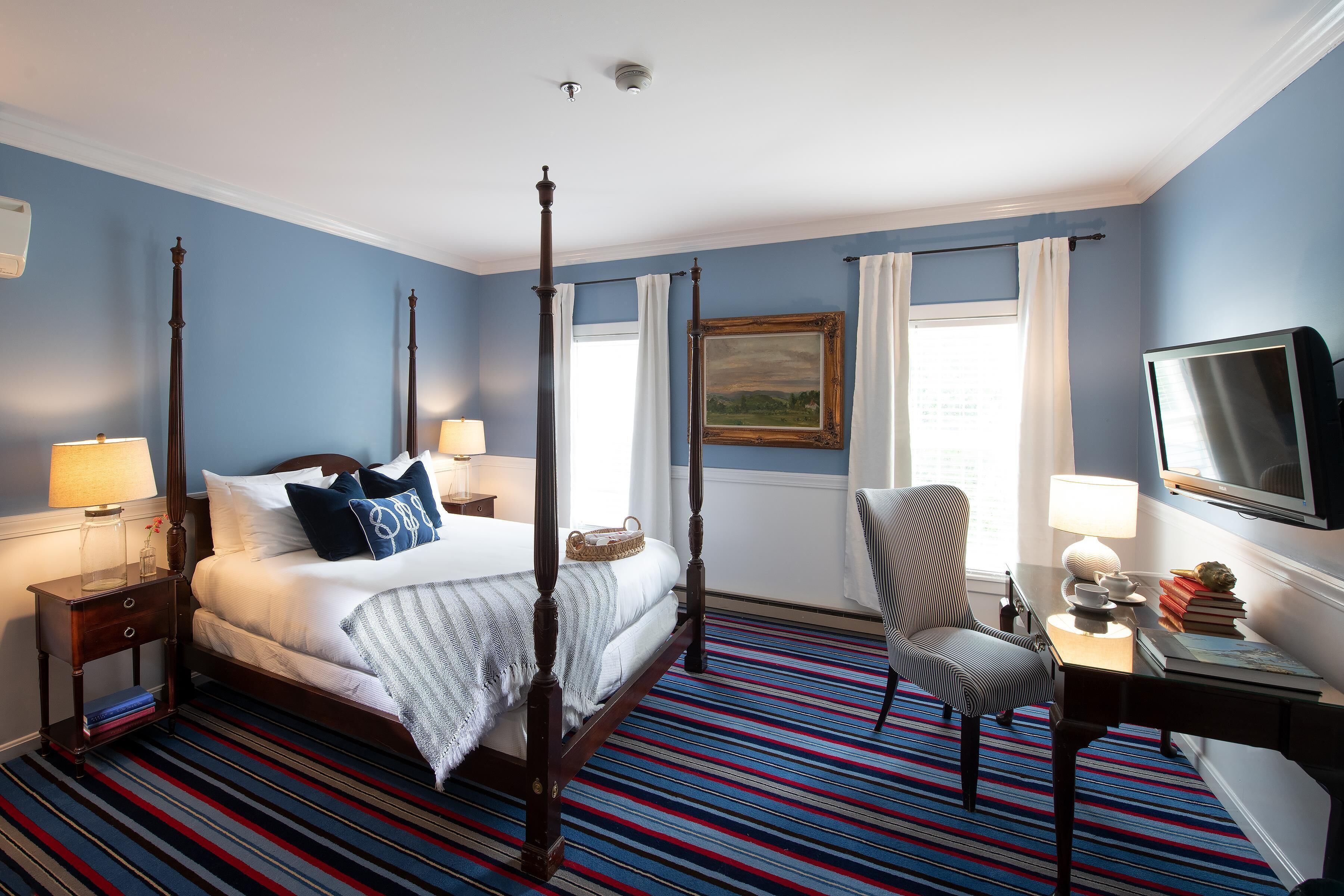 A four-poster bed with a white comforter and blue pillows, a striped carpet, a desk with a lamp, a chair, and a television.