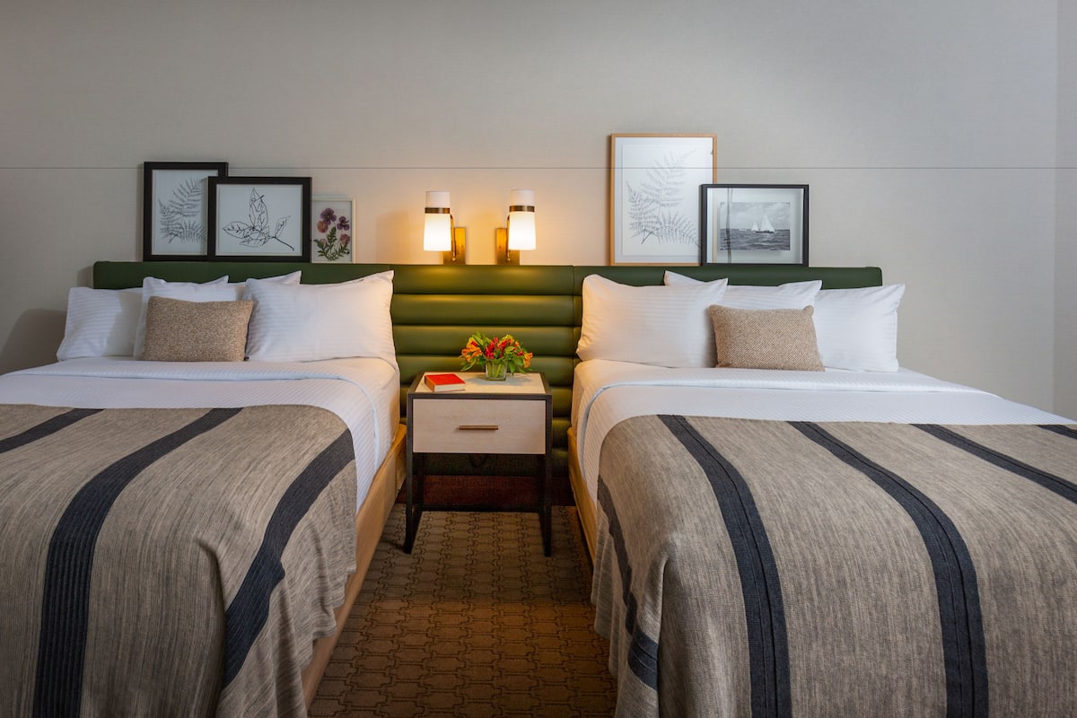 A hotel room with two beds, a nightstand with a vase of flowers, and framed artwork on the headboard.