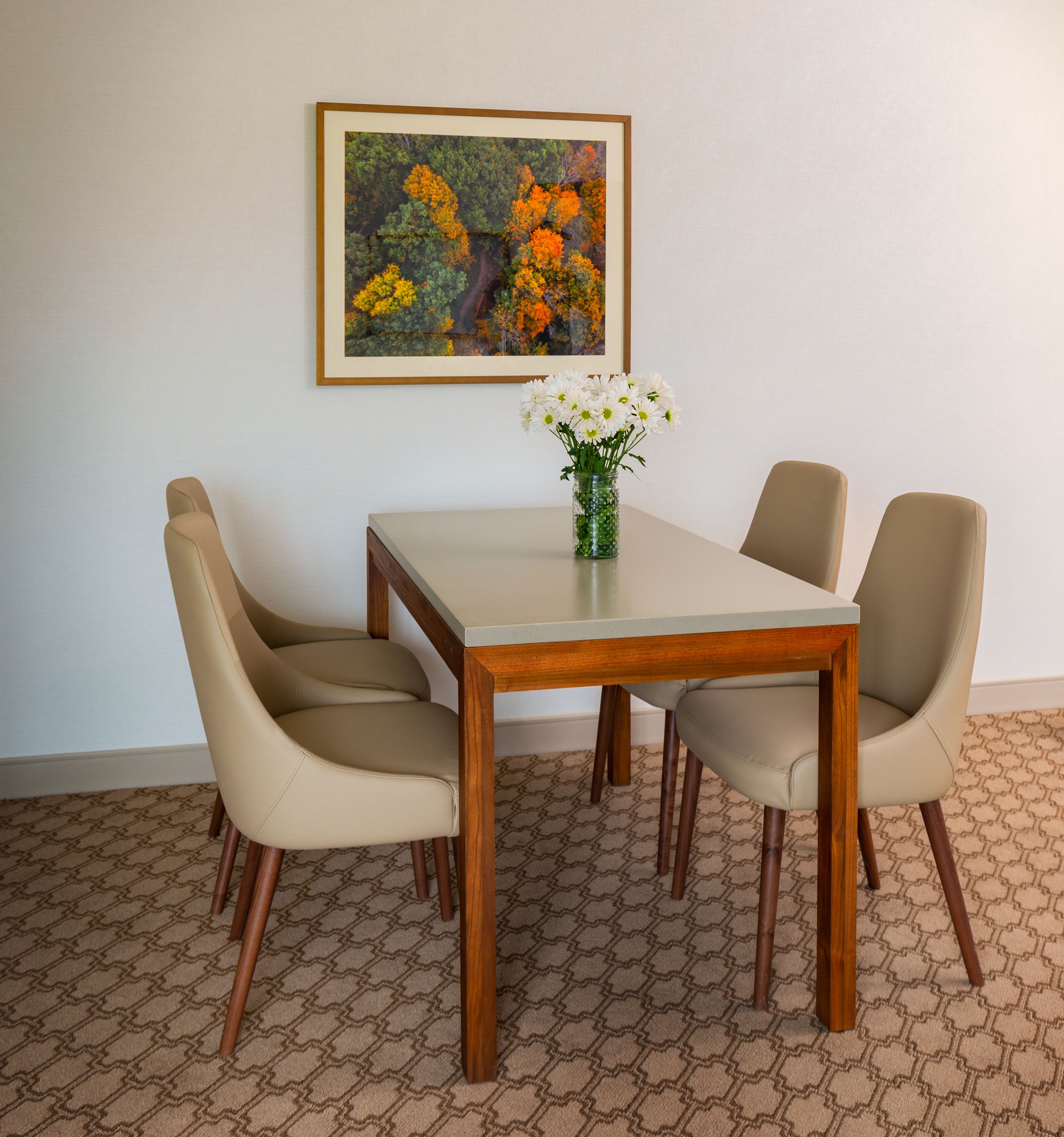 A dining area with a wooden table and four beige chairs. A framed painting of autumn foliage hangs on the wall.