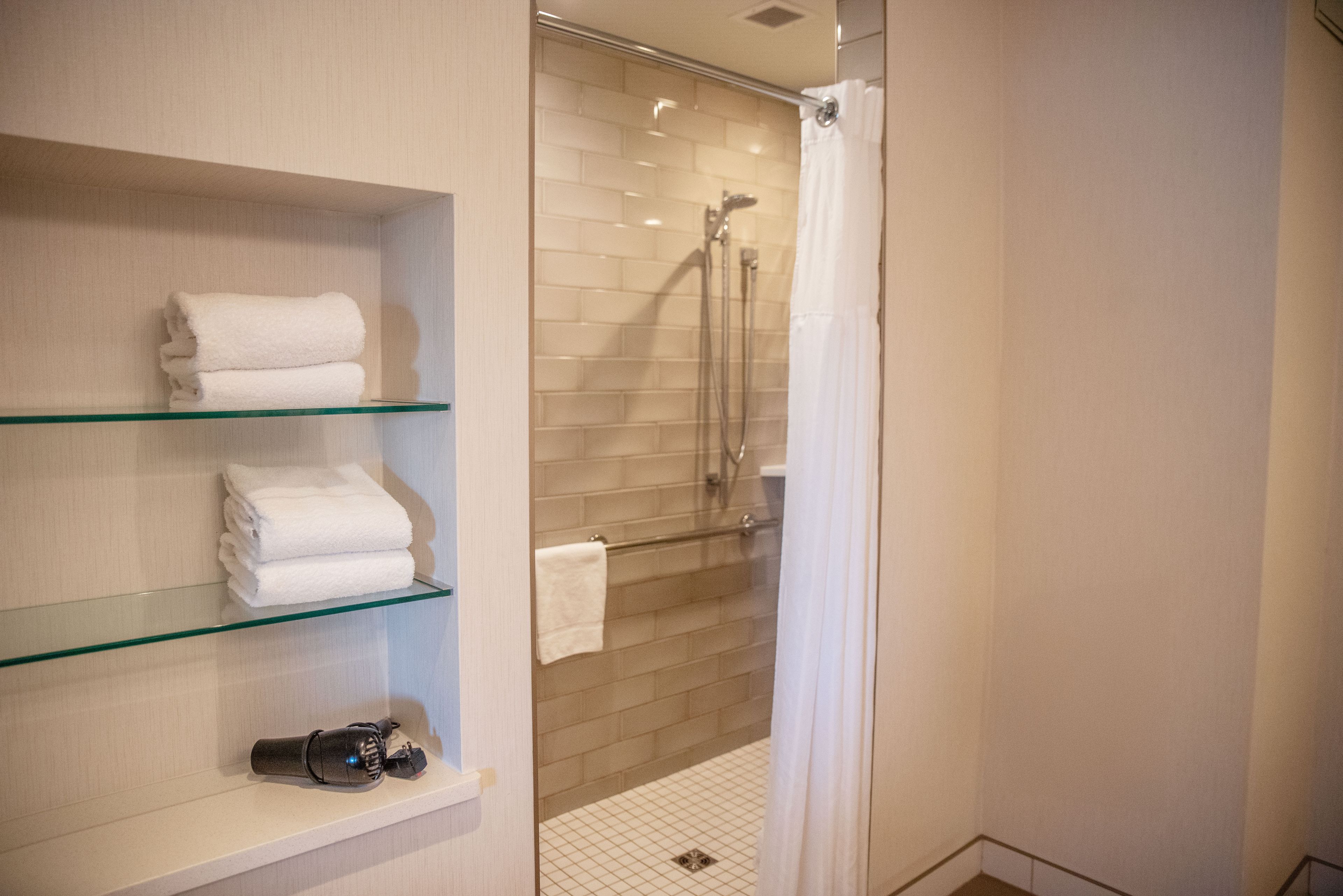 Un baño con repisas de vidrio que sostienen toallas dobladas, una ducha con puerta de vidrio y piso de baldosas.