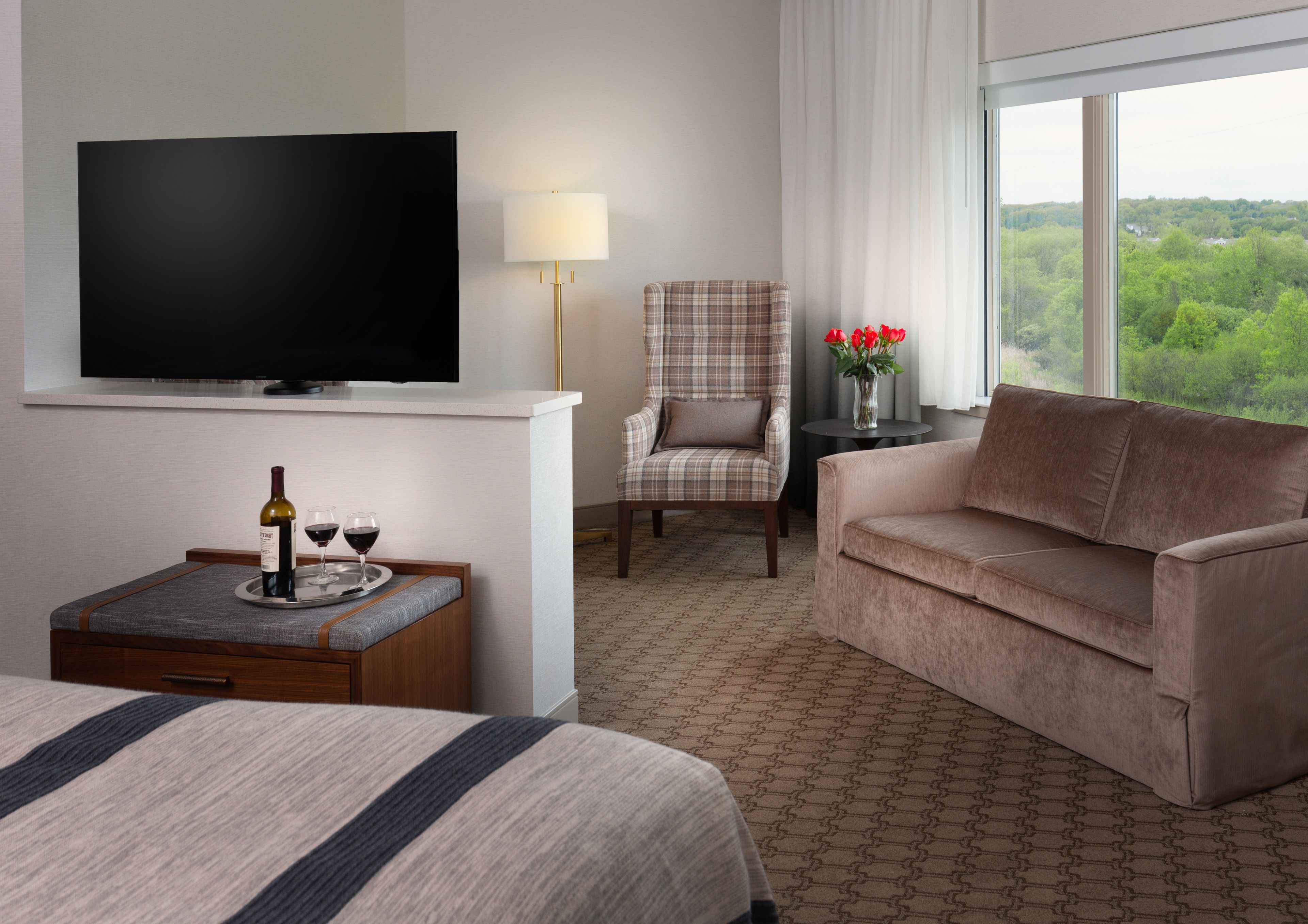 A hotel room with a bed, a TV, a sofa, a chair, a small table with a tray of wine glasses and a bottle, and a view of greenery through the window.
