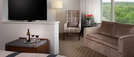 A hotel room with a bed, a TV, a sofa, a chair, a small table with a tray of wine glasses and a bottle, and a view of greenery through the window.