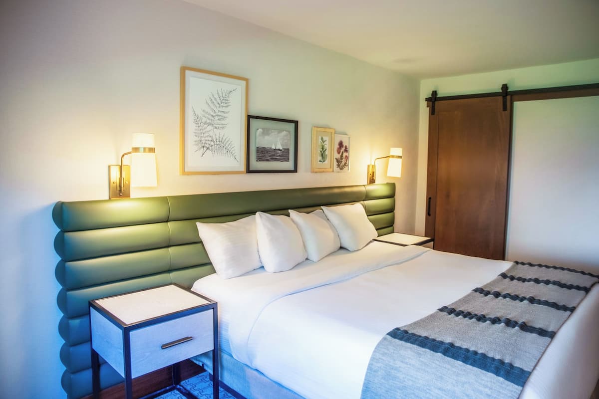 A hotel room with a bed, bedside tables, a green upholstered headboard, framed artwork on the wall, and a sliding door.