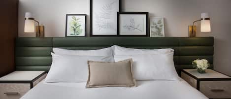 Habitación de hotel con una cama grande, cabecera tapizada en verde, ropa de cama blanca e impresiones botánicas enmarcadas en la pared.