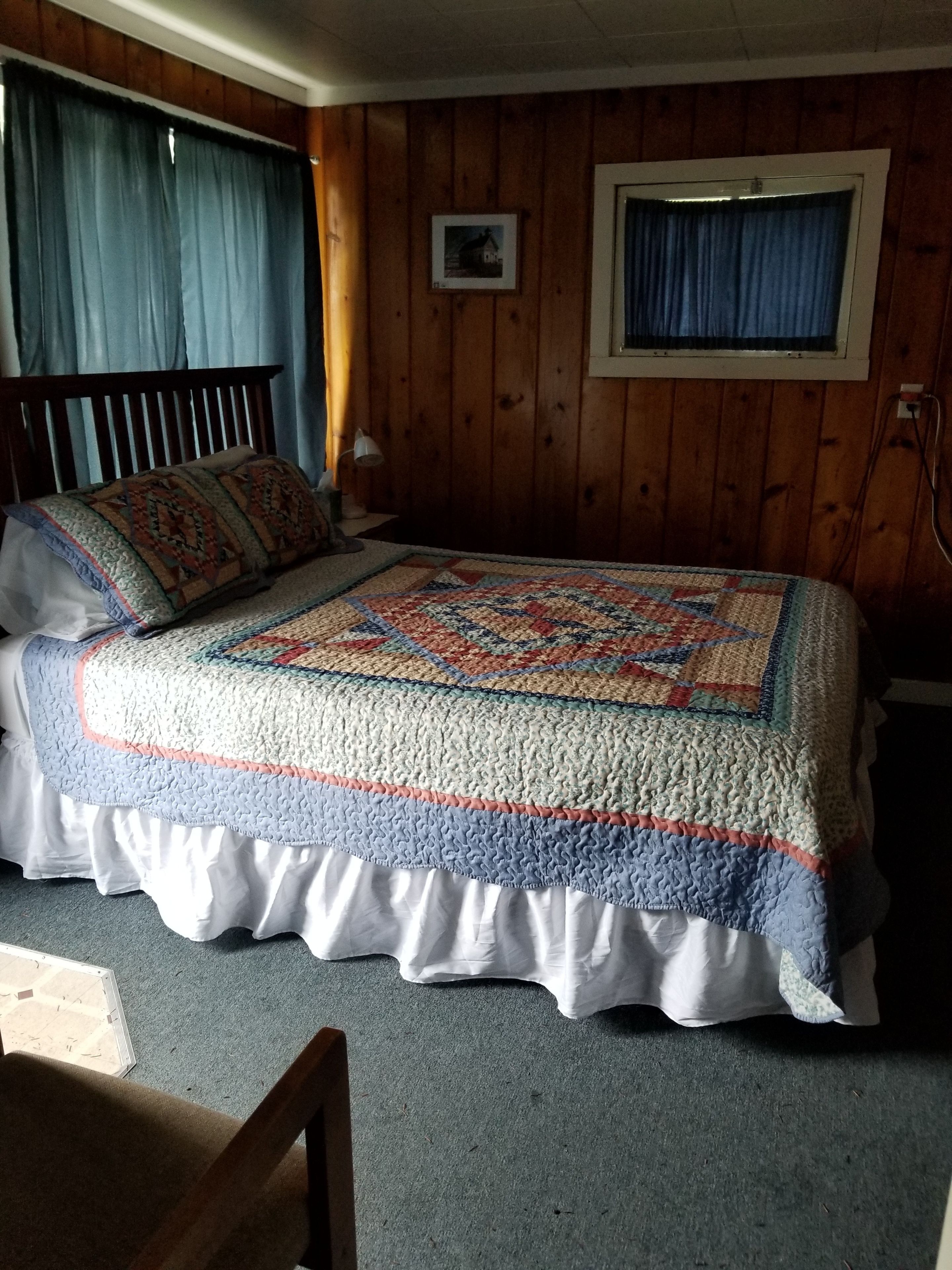Une chambre avec un mur lambrissé en bois, une couvre-lit matelassée, une lampe de chevet et un tableau encadré accroché au mur.