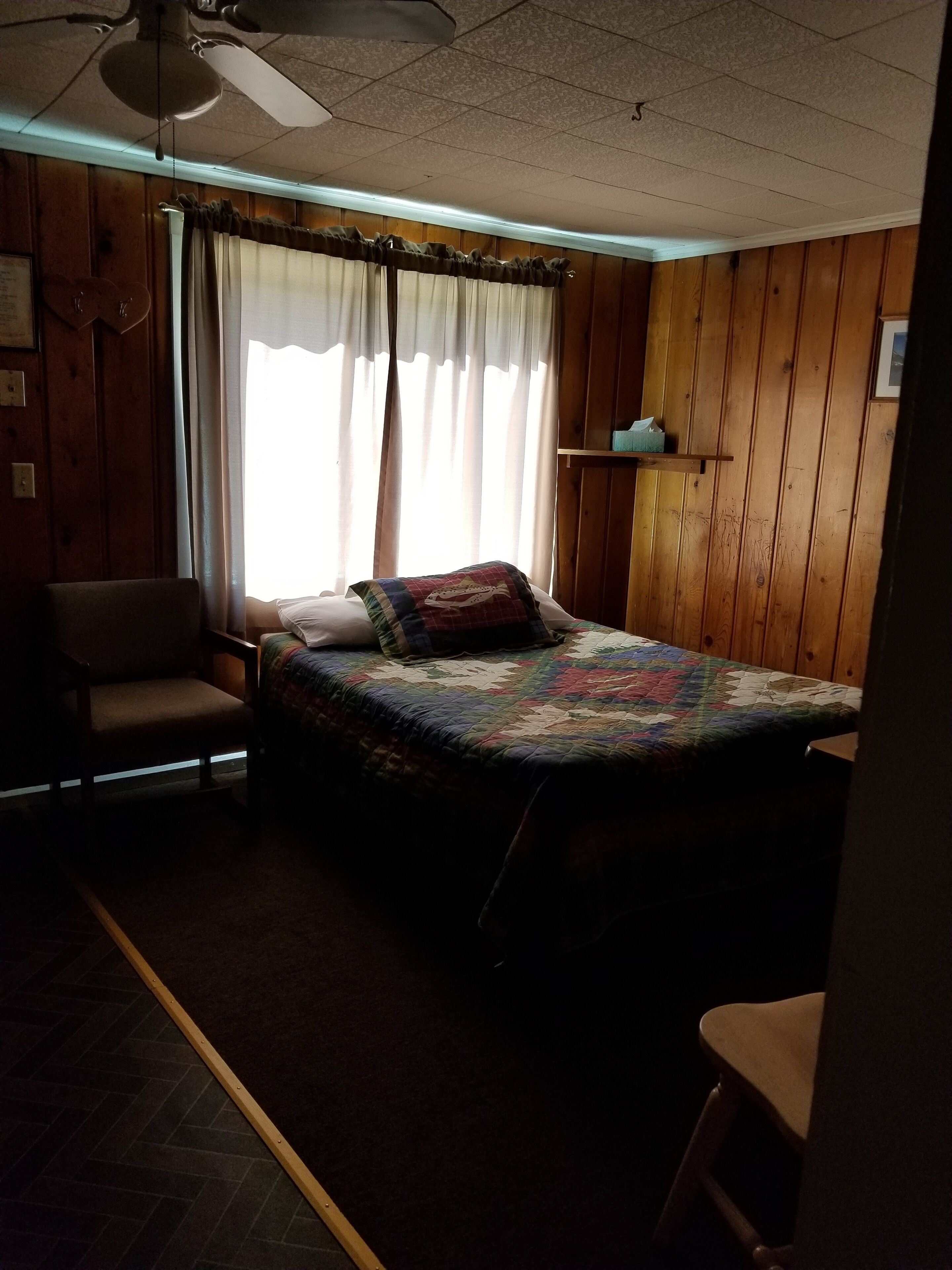 Una stanza con un letto, una sedia, una ventola a soffitto, pareti in legno, una finestra con tende e un piccolo ripiano.