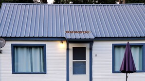A single-story house with a blue roof, a front door, and a small porch area. There is a white outdoor table and chairs, and a purple umbrella.