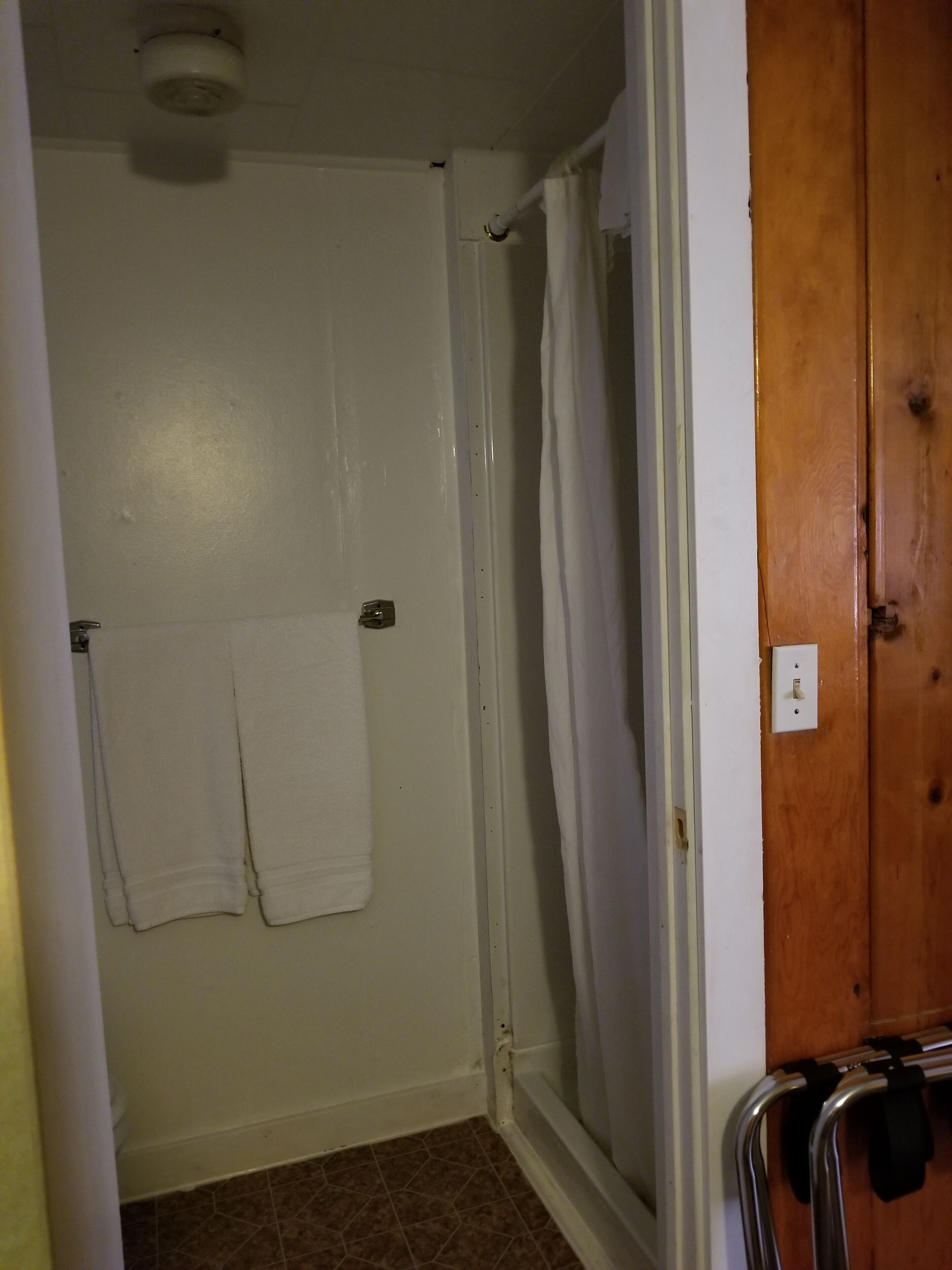 Un baño con ducha, un perchero con dos toallas, una puerta de madera y un interruptor de luz.