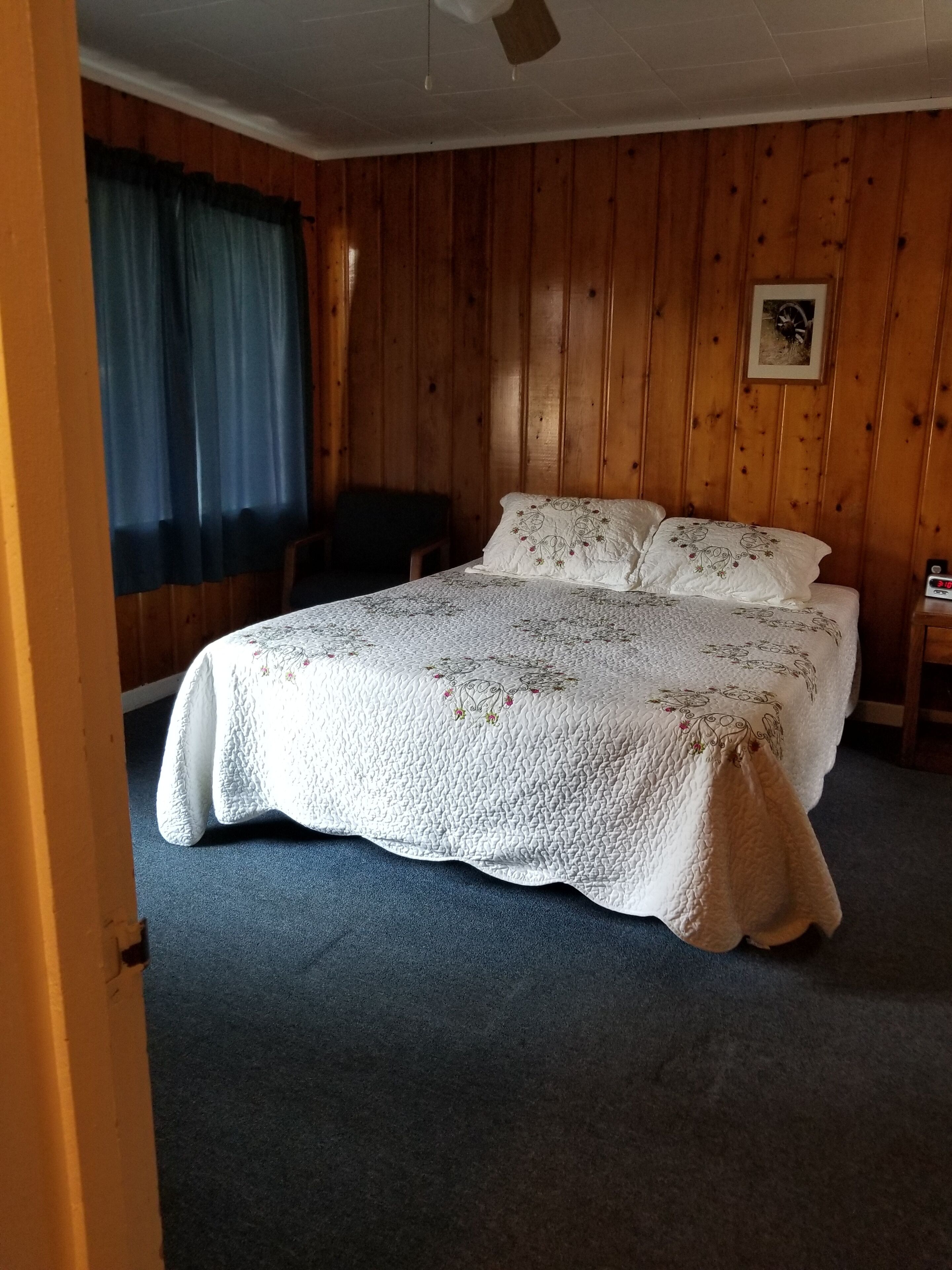 Una habitación con cama, una silla, paredes de madera, ventilador de techo y un cuadro en la pared.