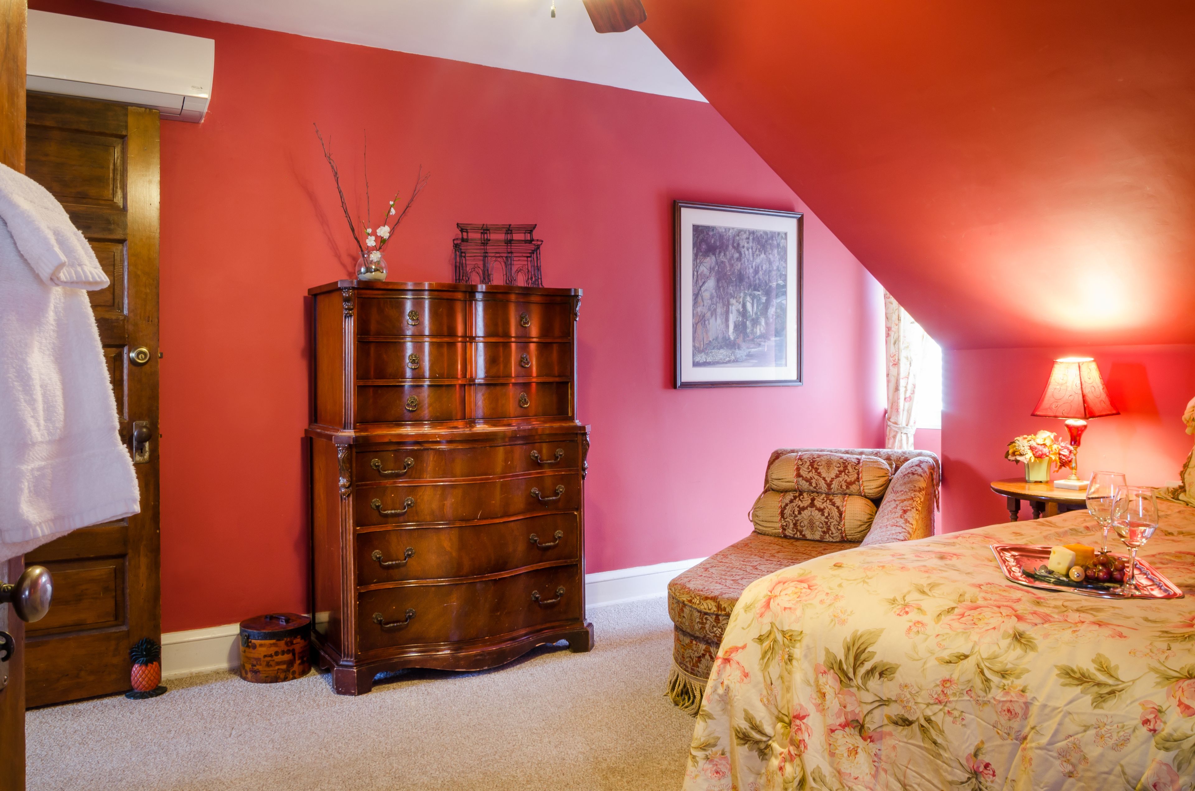 Ein Schlafzimmer mit roter Wand, einem hölzernen Schminktisch, einer blumigen Tagesdecke, einer roten Lampe und einem gerahmten Bild an der Wand.