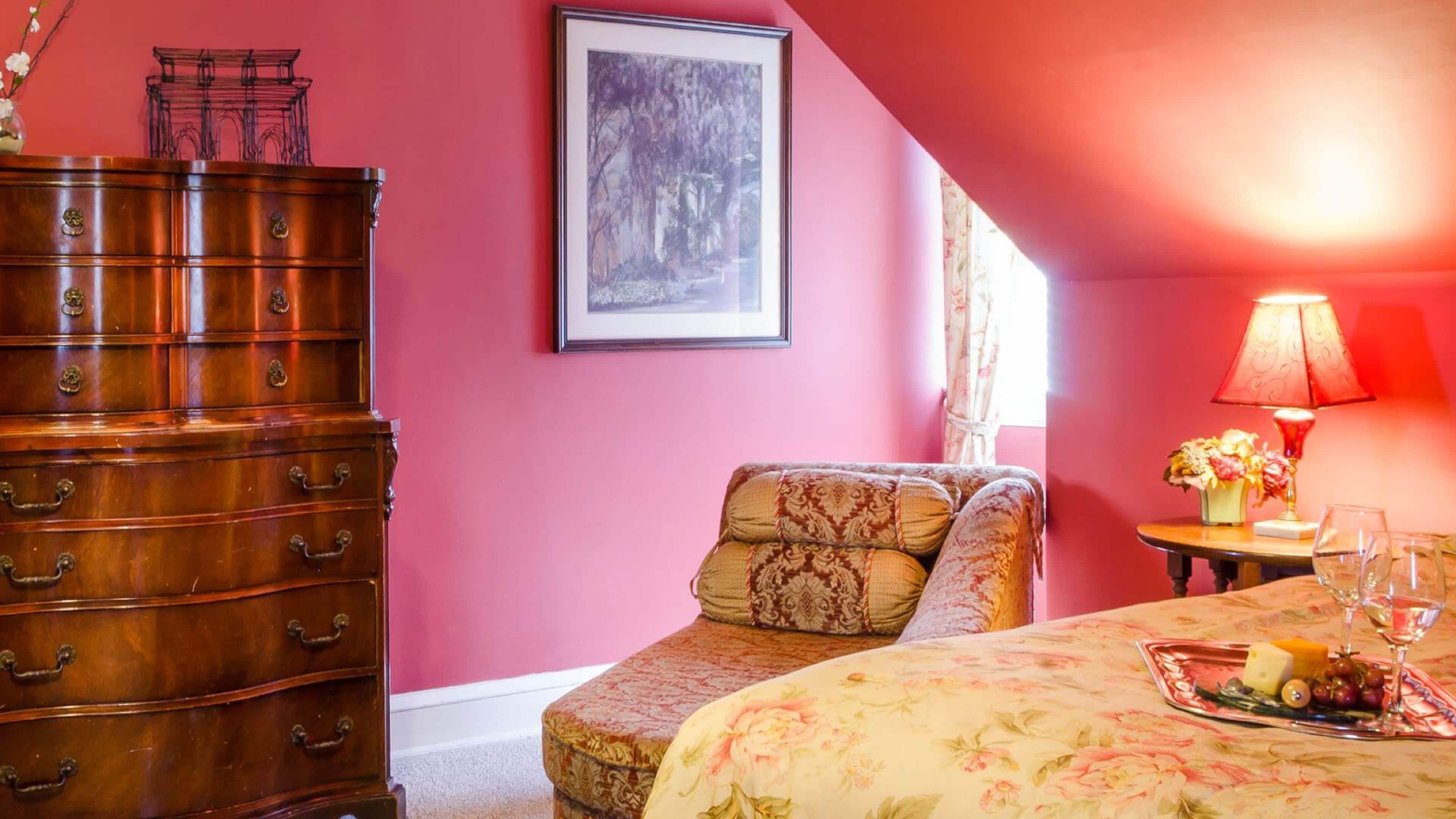 A room with a wooden dresser, a patterned armchair, a bed with a floral bedspread, a framed picture on the wall, and a table with a lamp and flowers.