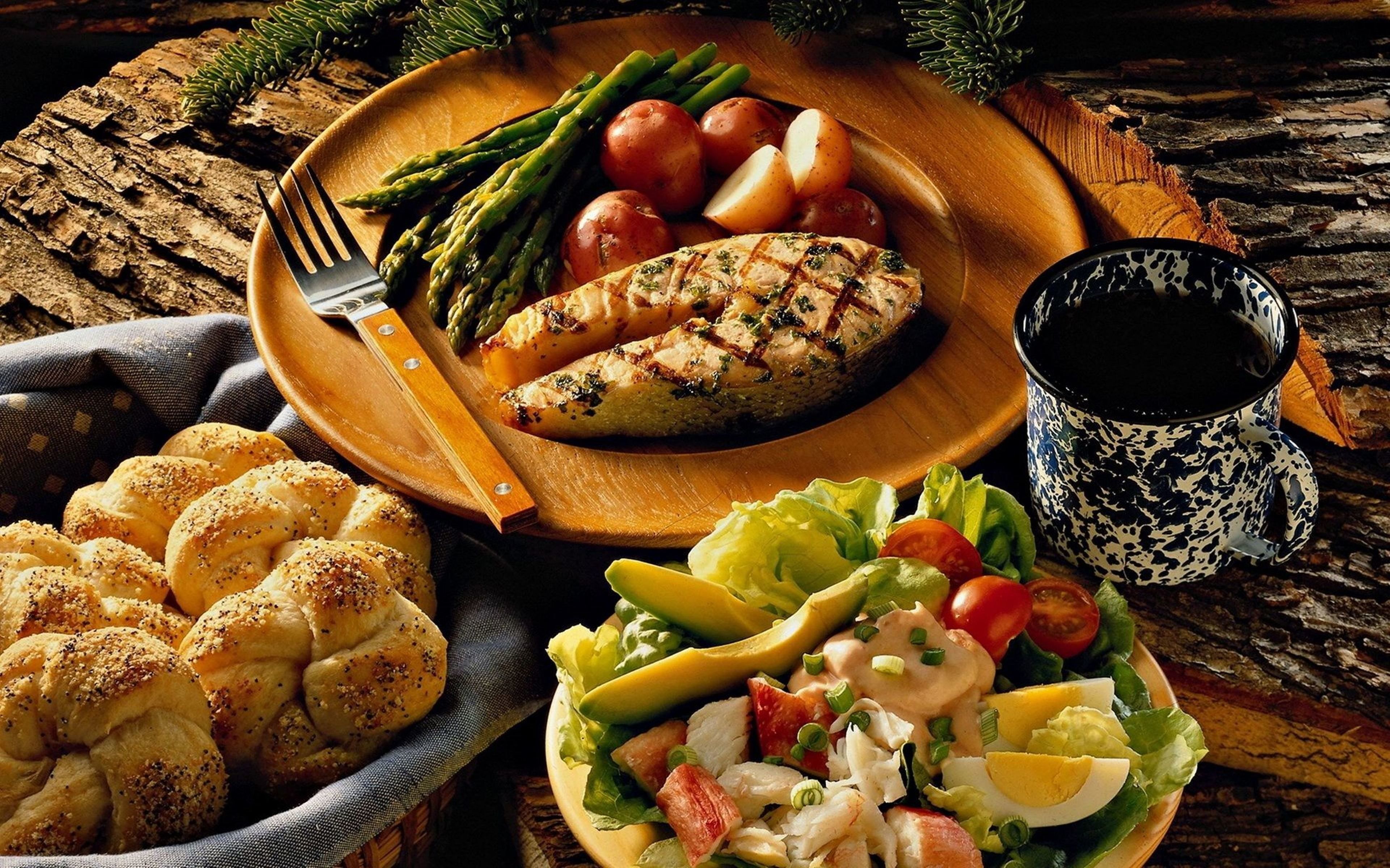 A plate with grilled chicken, asparagus, and potatoes, a bowl of bread rolls, a salad with avocado and eggs, and a cup of coffee on a rustic wooden table.
