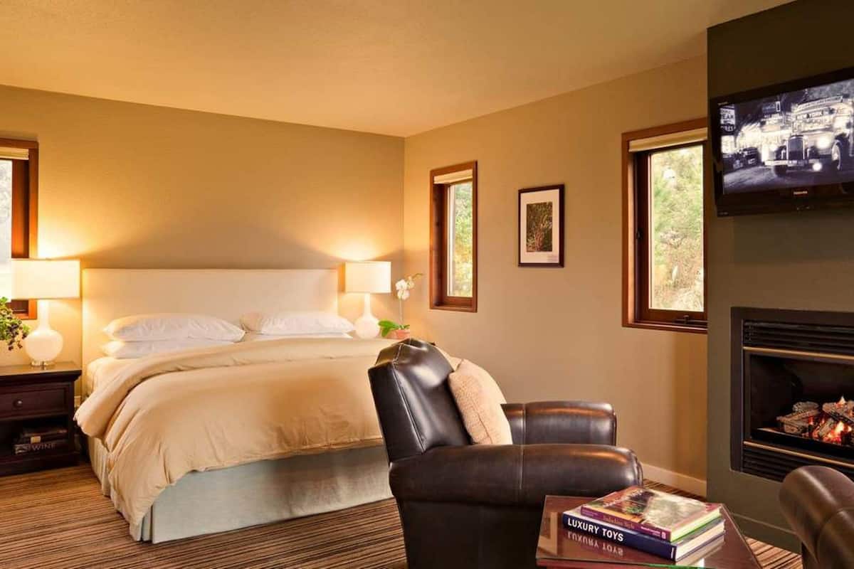 A hotel room with a bed, a leather chair, a fireplace, a television, and a nightstand with a lamp and a vase of flowers.