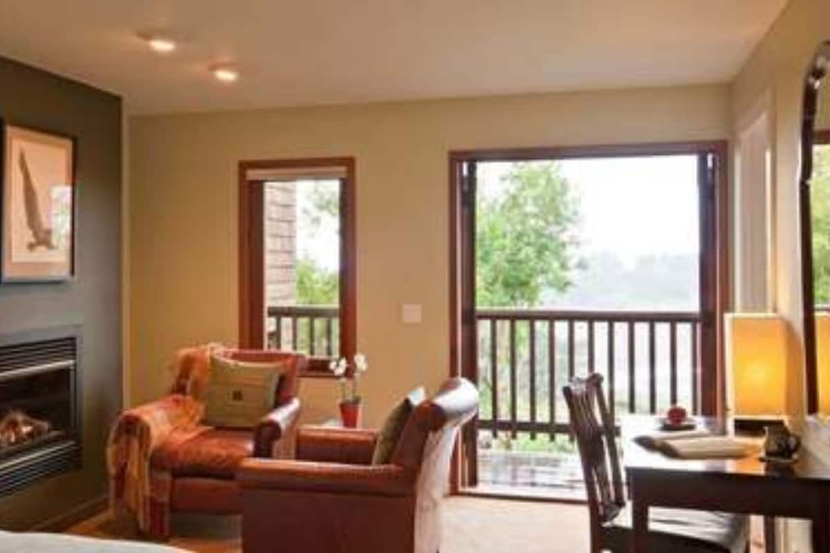 A room with a fireplace, a sofa, a desk, and a balcony with a view of greenery.