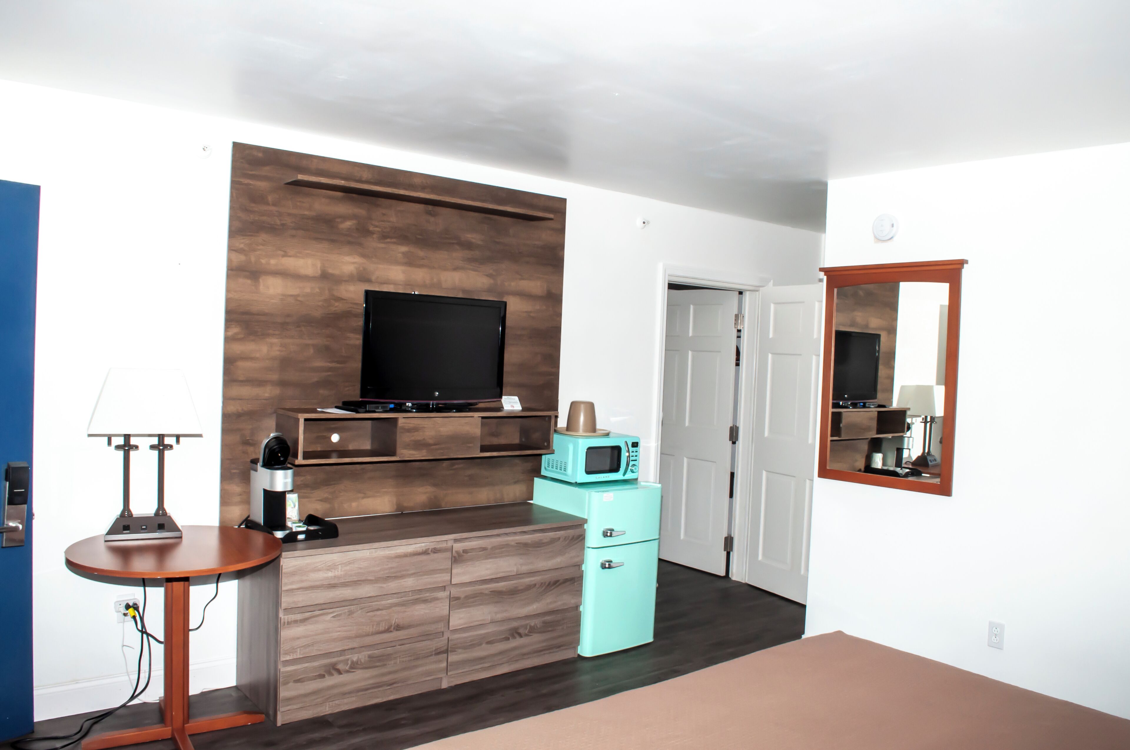 A modern living room with a flat-screen TV, a coffee maker, a microwave, and a small fridge. There is a wooden cabinet and a round table with a lamp.