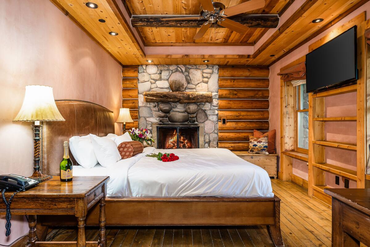 A cozy log cabin room with a stone fireplace, wooden ceiling, and a bed with white linens.