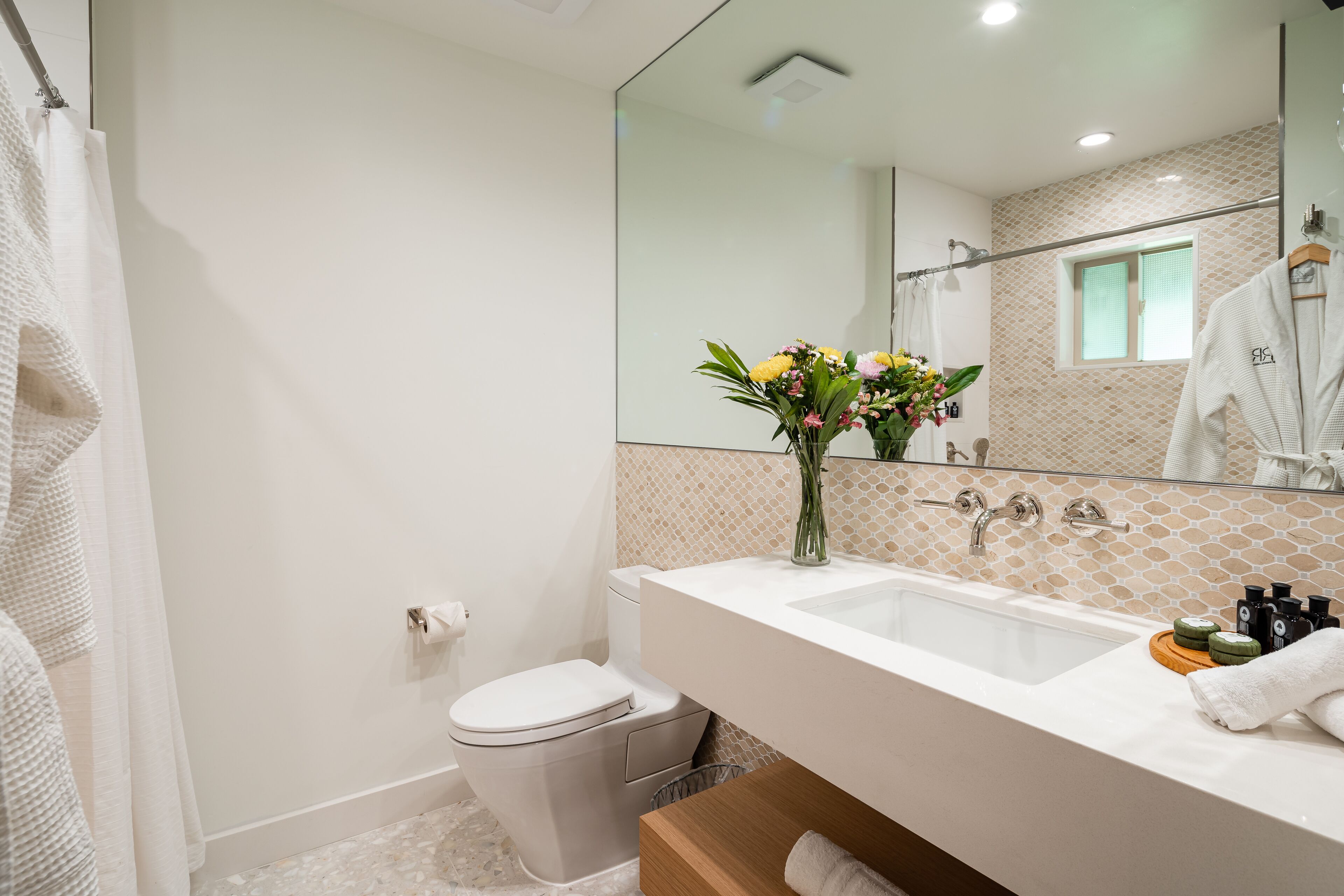 Un baño moderno con lavamanos, espejo y váter blancos. Hay un florero con flores sobre el mostrador del lavamanos.