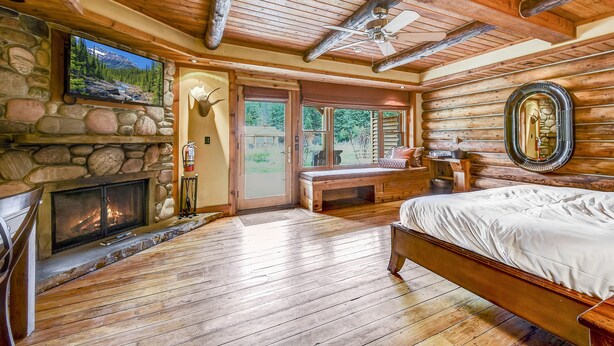 A log cabin interior with a stone fireplace, a bed, a bench, and a television mounted on the wall. - Rainbow Ranch Lodge (Gallatin Gateway)