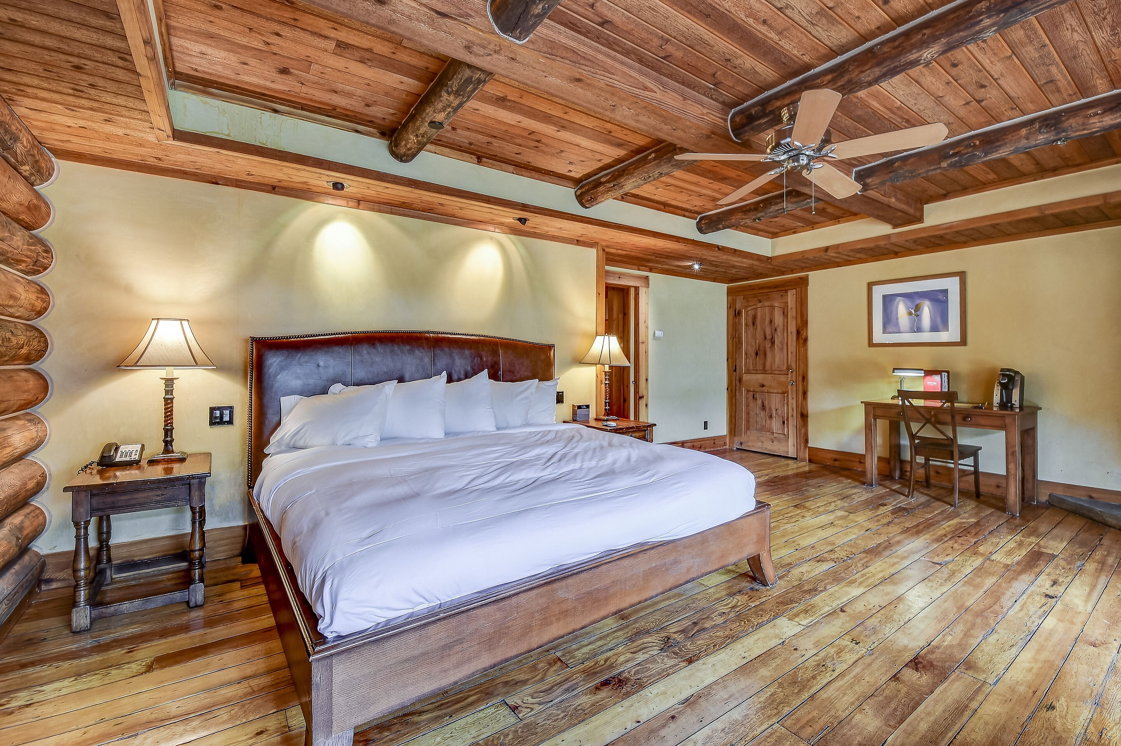 Un dormitorio de estilo cabaña de madera con una cama de madera, dos mesitas de noche con lámparas, un escritorio con una silla y un ventilador de techo.