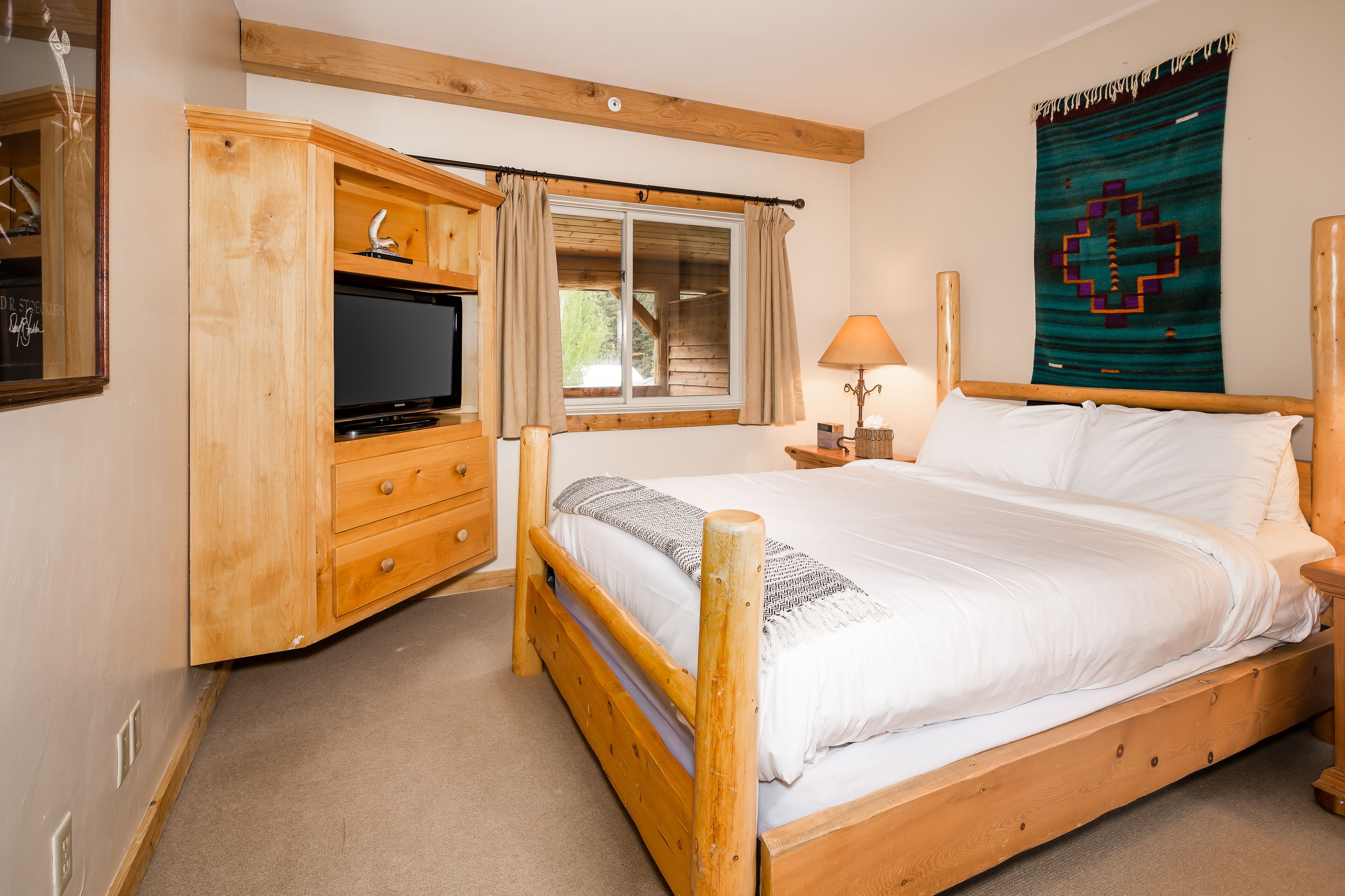 A wooden bed with white linens, a wooden nightstand with a lamp, a wooden wardrobe, and a window with curtains.