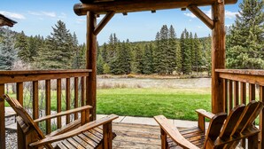 A wooden deck with two chairs facing a grassy area and a forest backdrop. - Rainbow Ranch Lodge (Gallatin Gateway)
