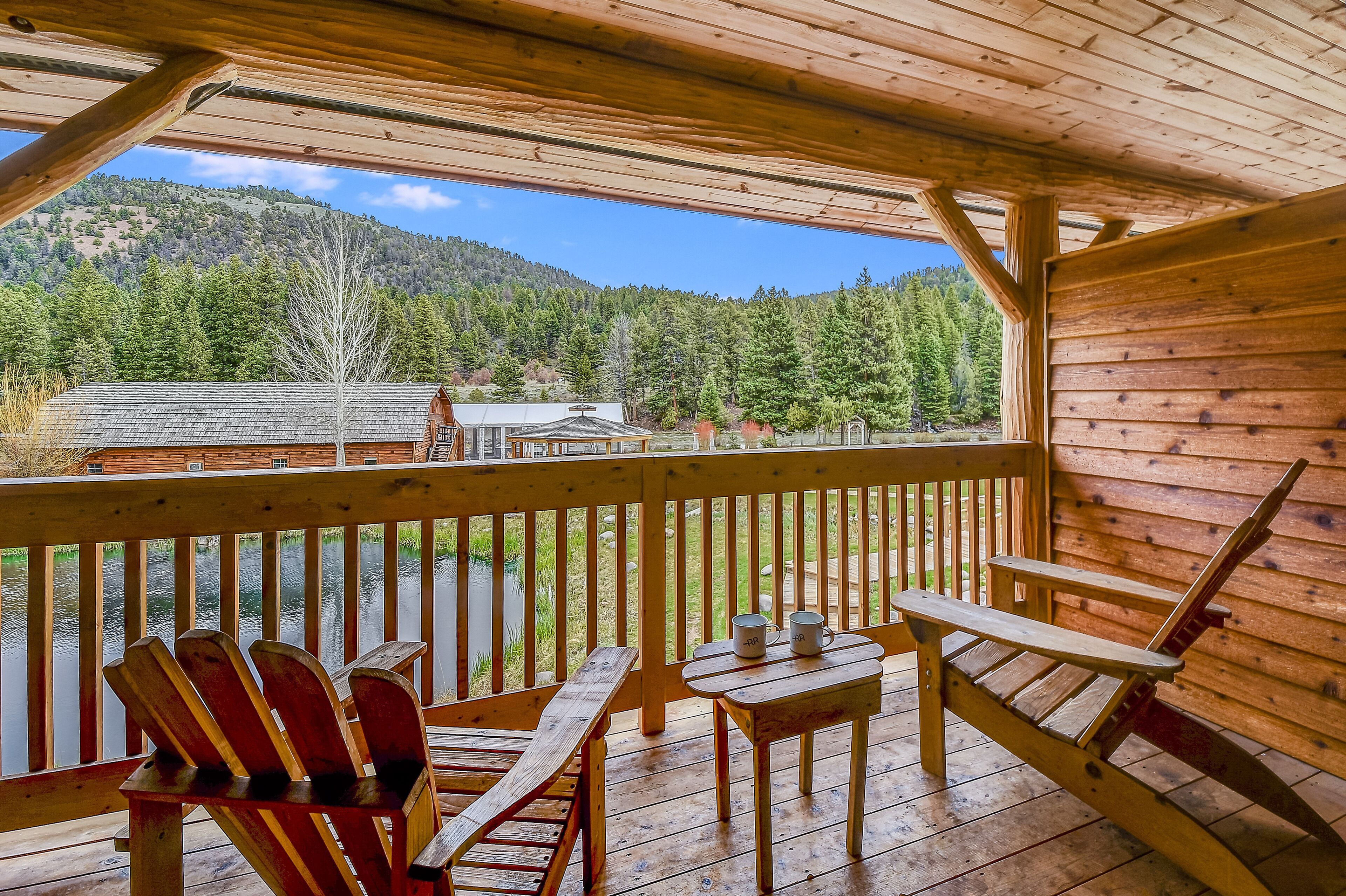 Una terraza de madera con sillas Adirondack y una mesa, con vista a un lago y montañas boscosas.