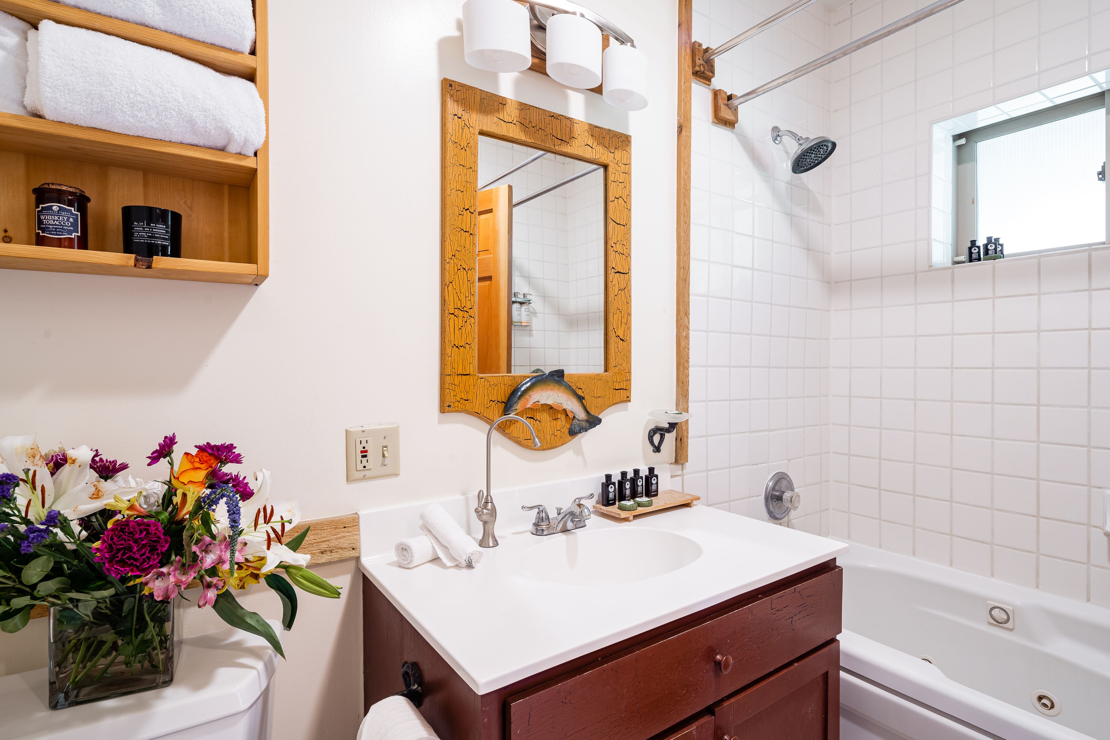 Un baño con tocador de madera, lavabo blanco, espejo, ducha y estante con toallas y artículos de aseo.