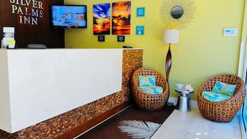 Un área de recepción con mostrador, dos sillas de mimbre con cojines, un televisor montado en la pared y un espejo decorativo con diseño de sol.