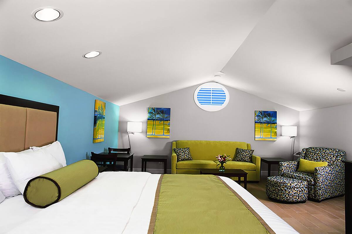 A hotel room with a bed, a green sofa, a patterned armchair, a small table with a vase, and two wall-mounted paintings.