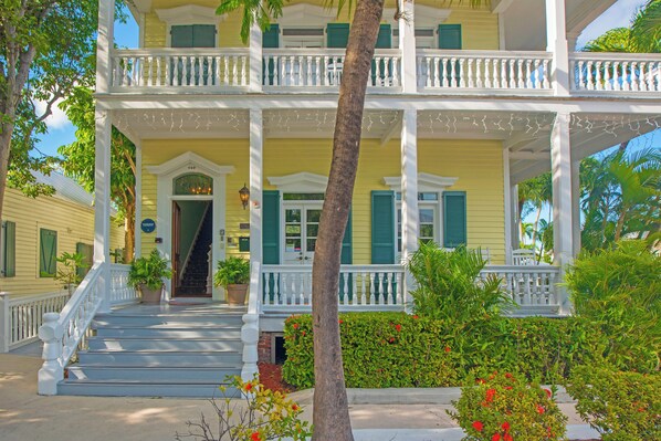 A two-story yellow house with a porch, white railings, and green shutters.